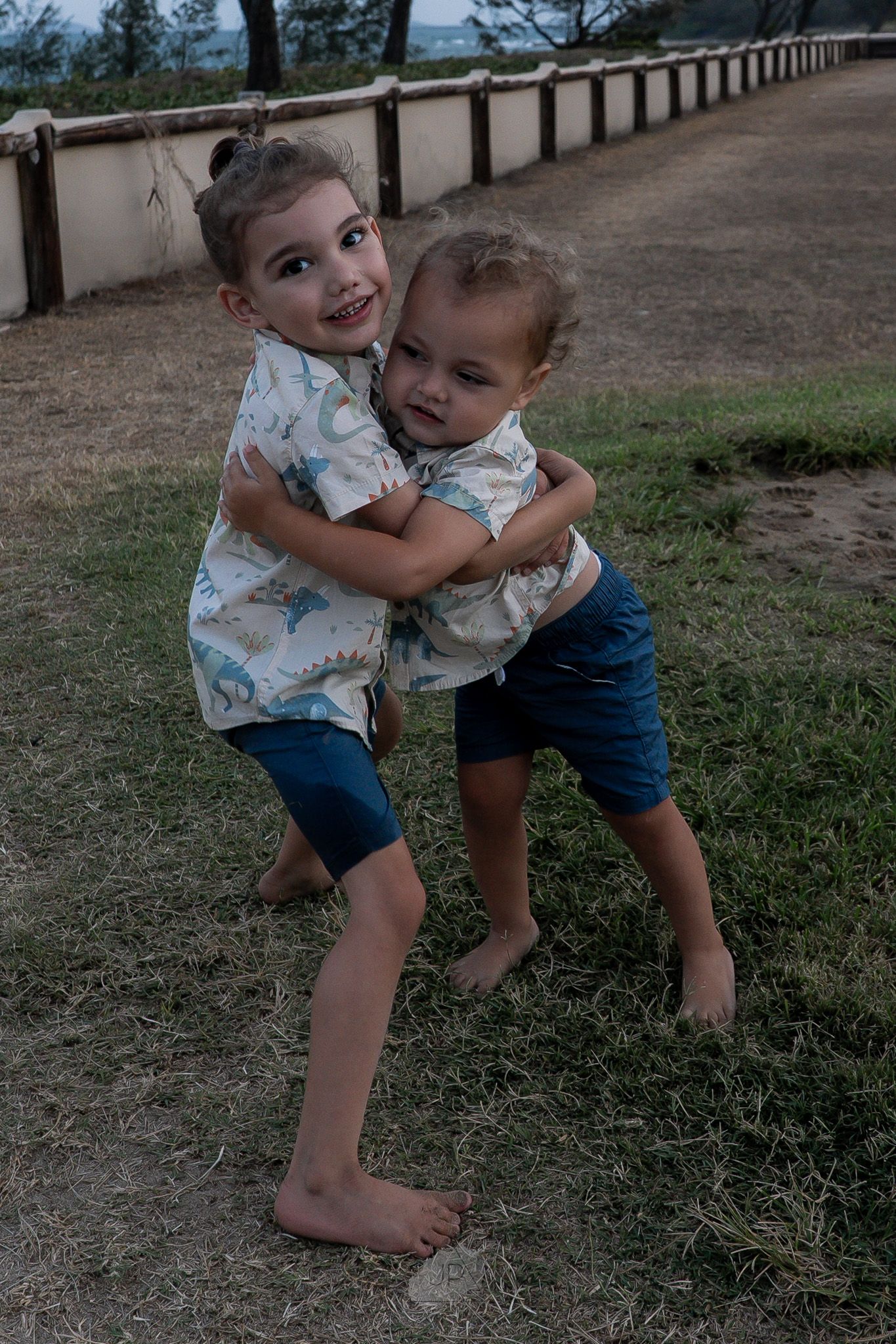 Two children playfully wrestling and hugging on a dirt path next to a railing during an outdoor photo session.