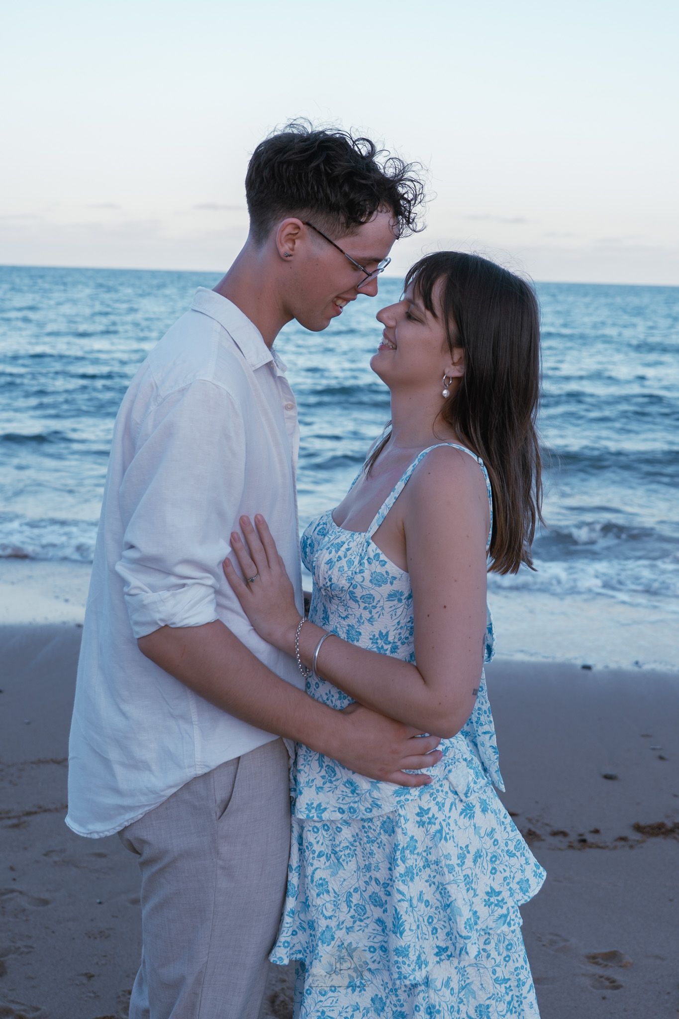A romantic couple embraces on a beach at sunset while wearing white and blue floral attire.