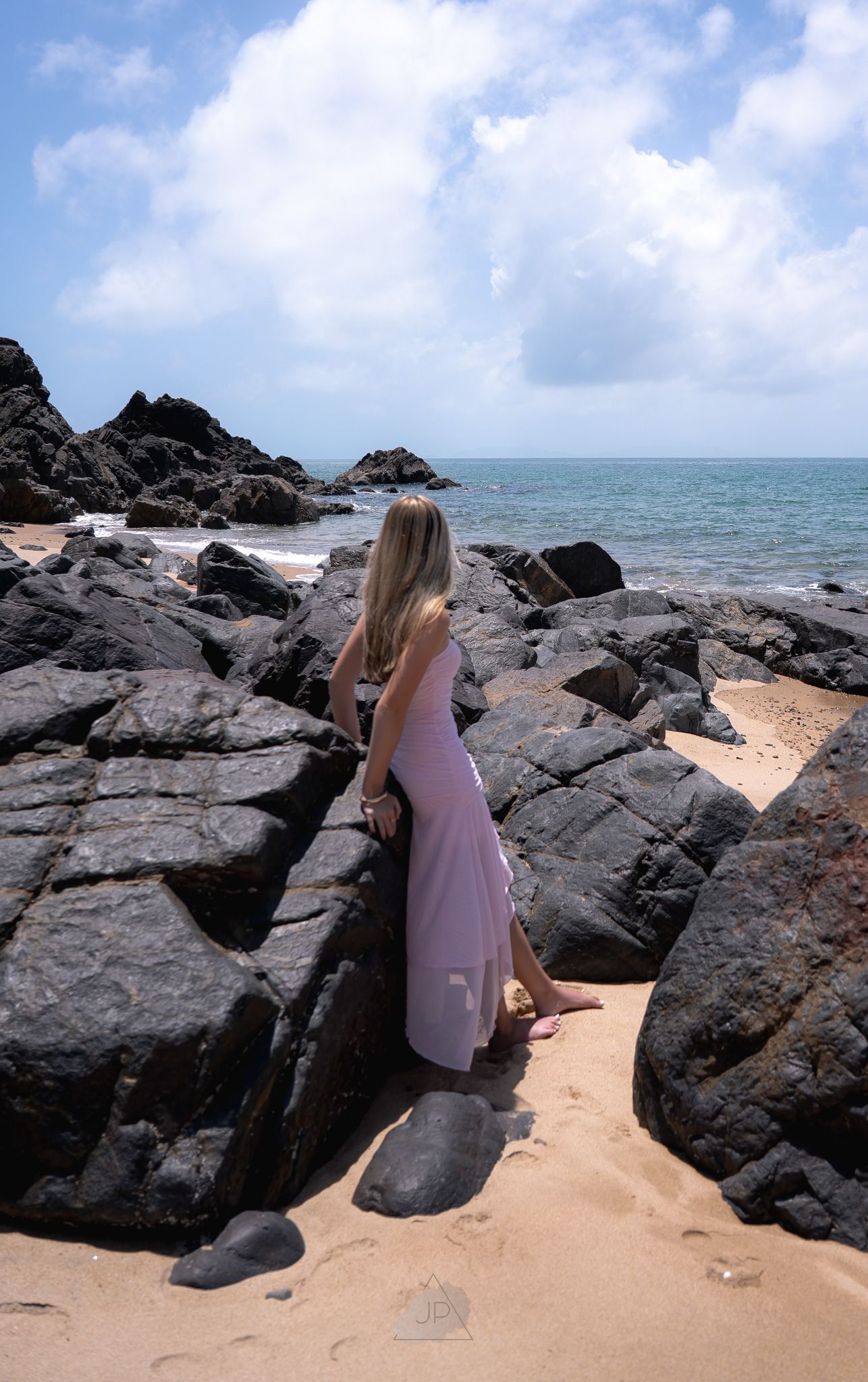A woman in a pink dress explores a rocky beach coastline with dark volcanic rocks and sandy patches near the ocean.