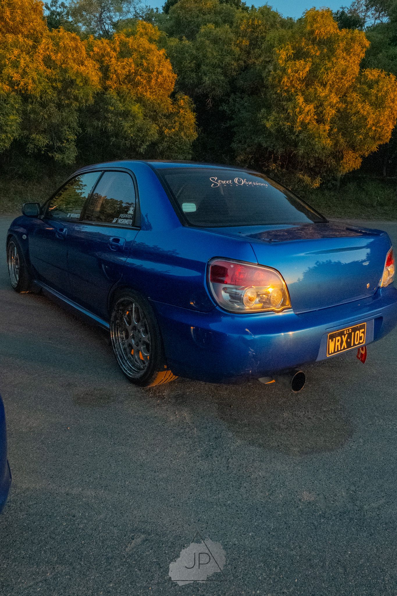 A modified blue Nissan Silvia S15 sports car shown at sunset with autumn trees in the background.