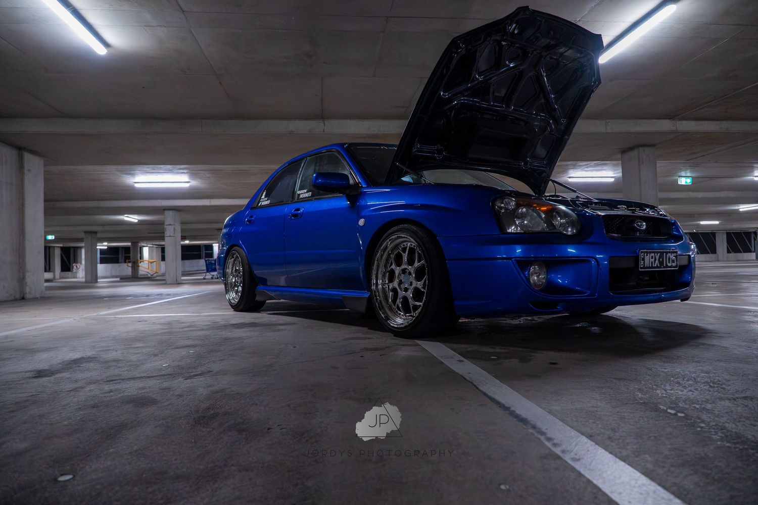 Blue Subaru WRX STI parked in a dimly lit concrete parking garage with fluorescent lighting overhead.