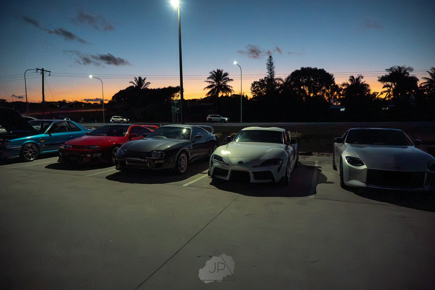 Luxury sports cars parked in a lot at dusk with palm trees and dramatic sunset lighting in the background.