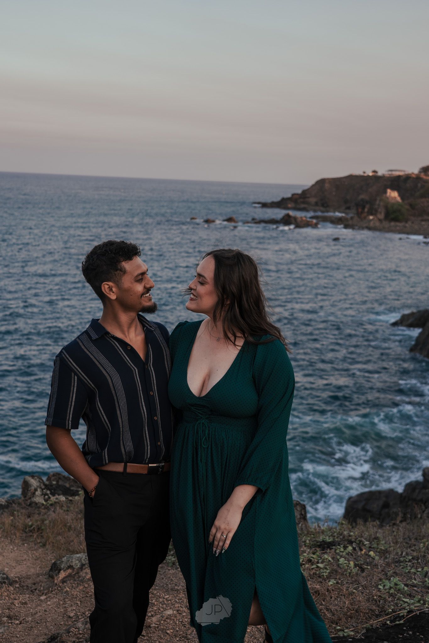 A couple shares intimate moments along a rocky coastal cliff at sunset with crashing waves in the background.