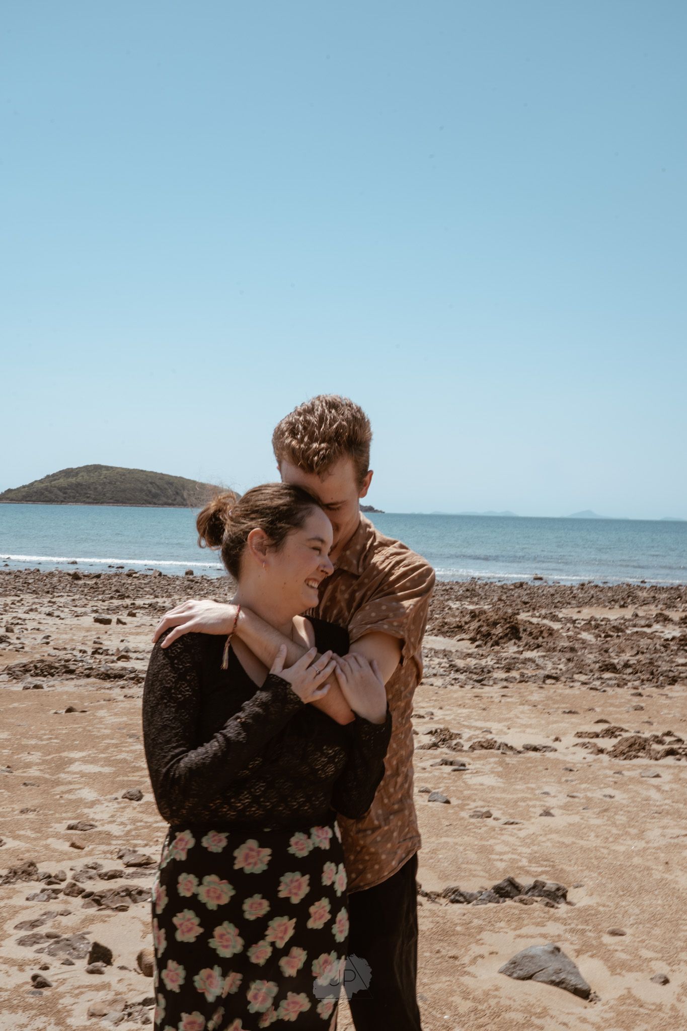 A couple shares a tender moment on a rocky beach against a blue sky and ocean backdrop.