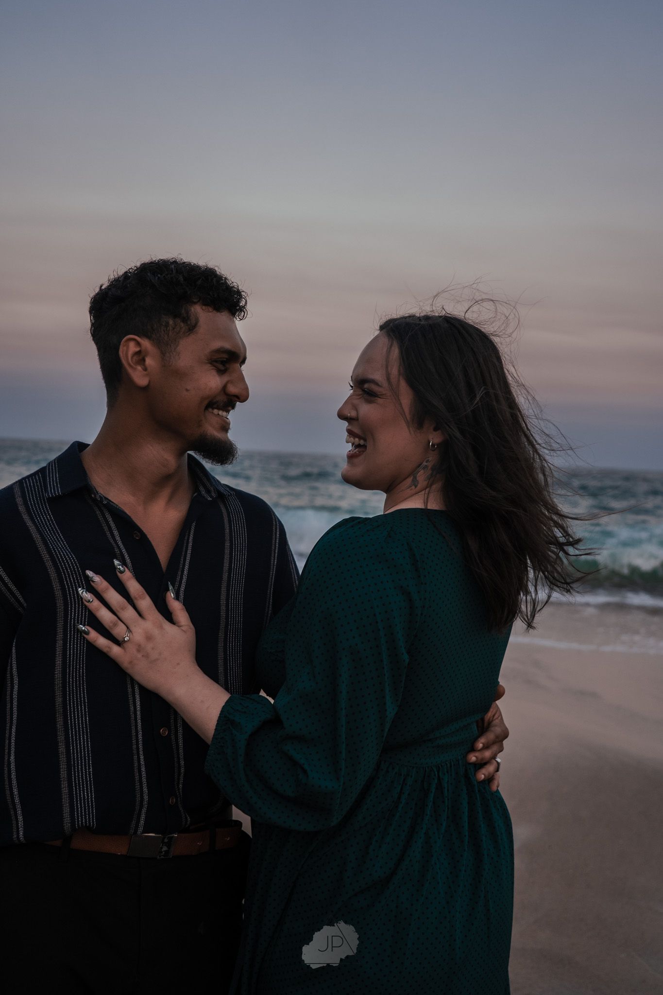A couple shares joyful laughter and embraces on a moody beach at sunset.