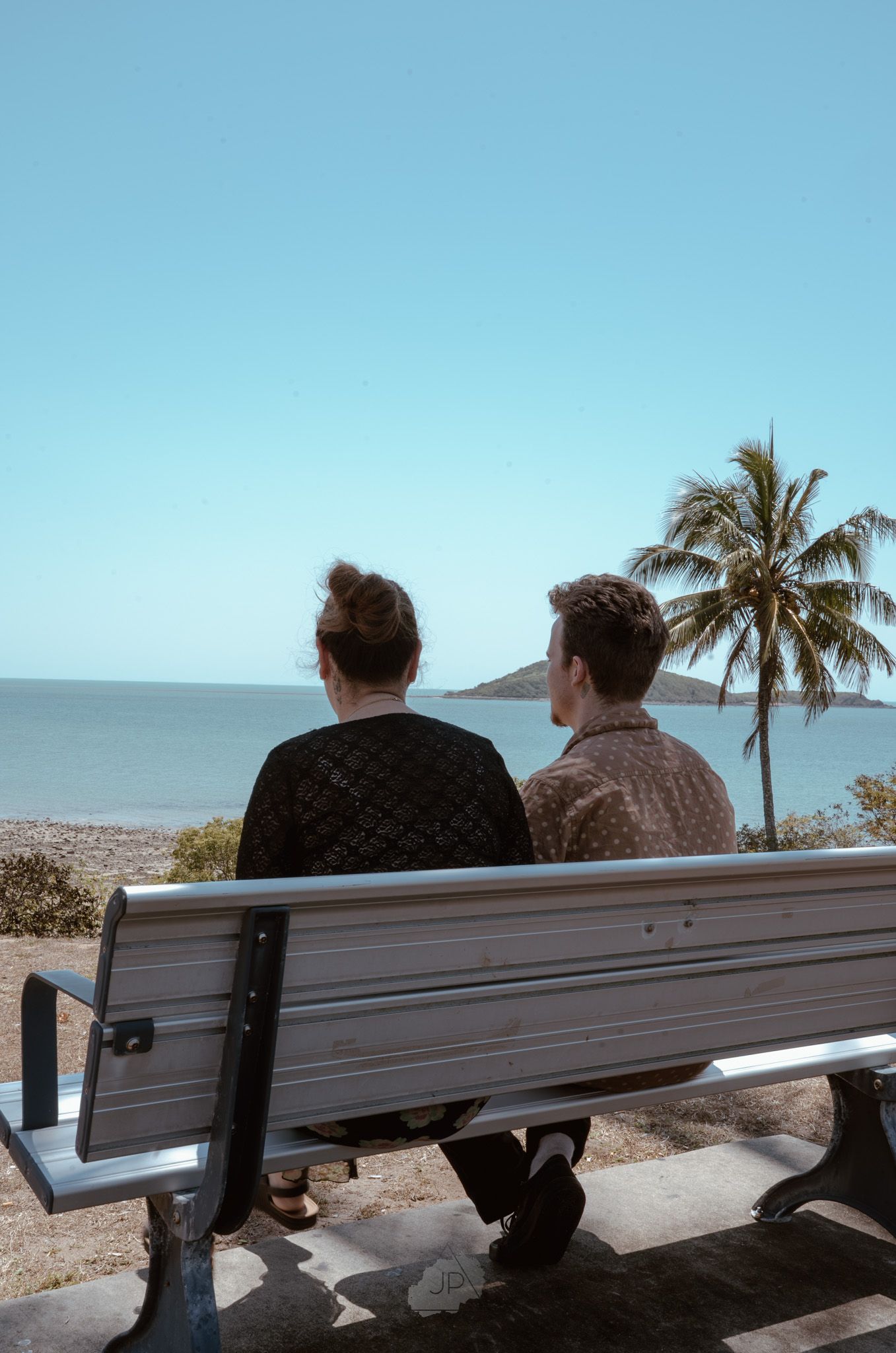 Two people sit together on a bench overlooking a desert landscape with palm trees and mountains in the distance.