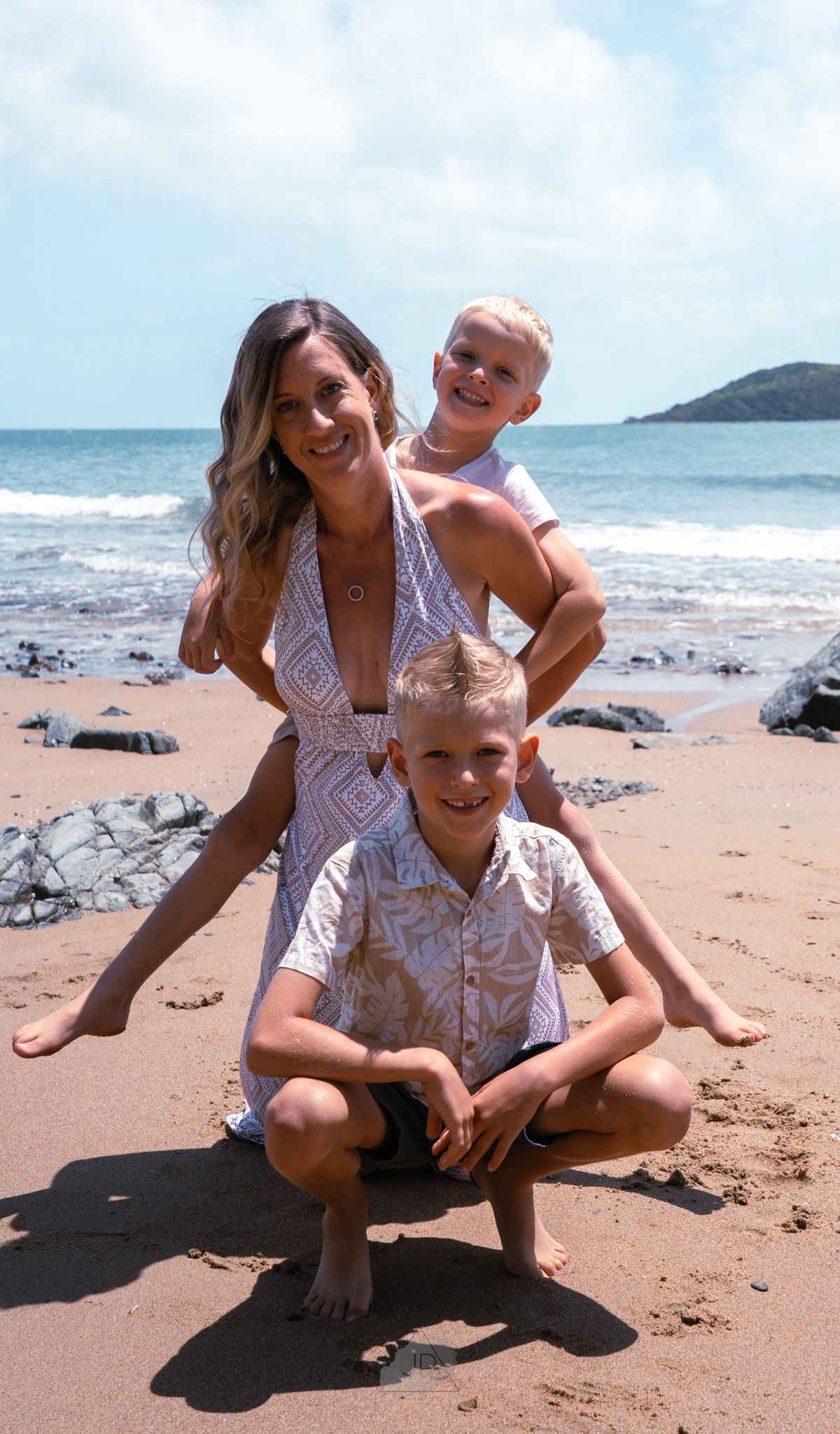 A family enjoys a sunny day at a rocky beach with blue ocean waters and mountains in the background.