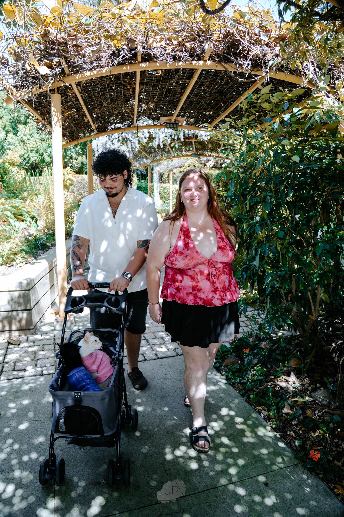 Two people sit together on a bench under a wooden pergola surrounded by lush garden foliage.