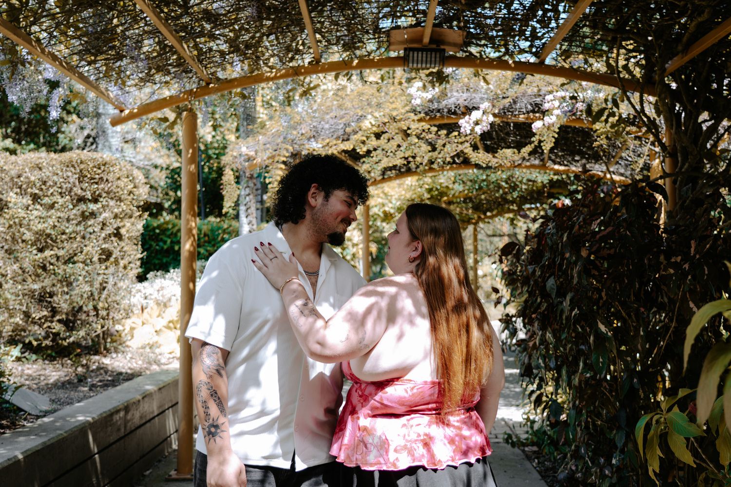 A romantic couple stands together under a flowering tree in a garden setting with dappled sunlight.