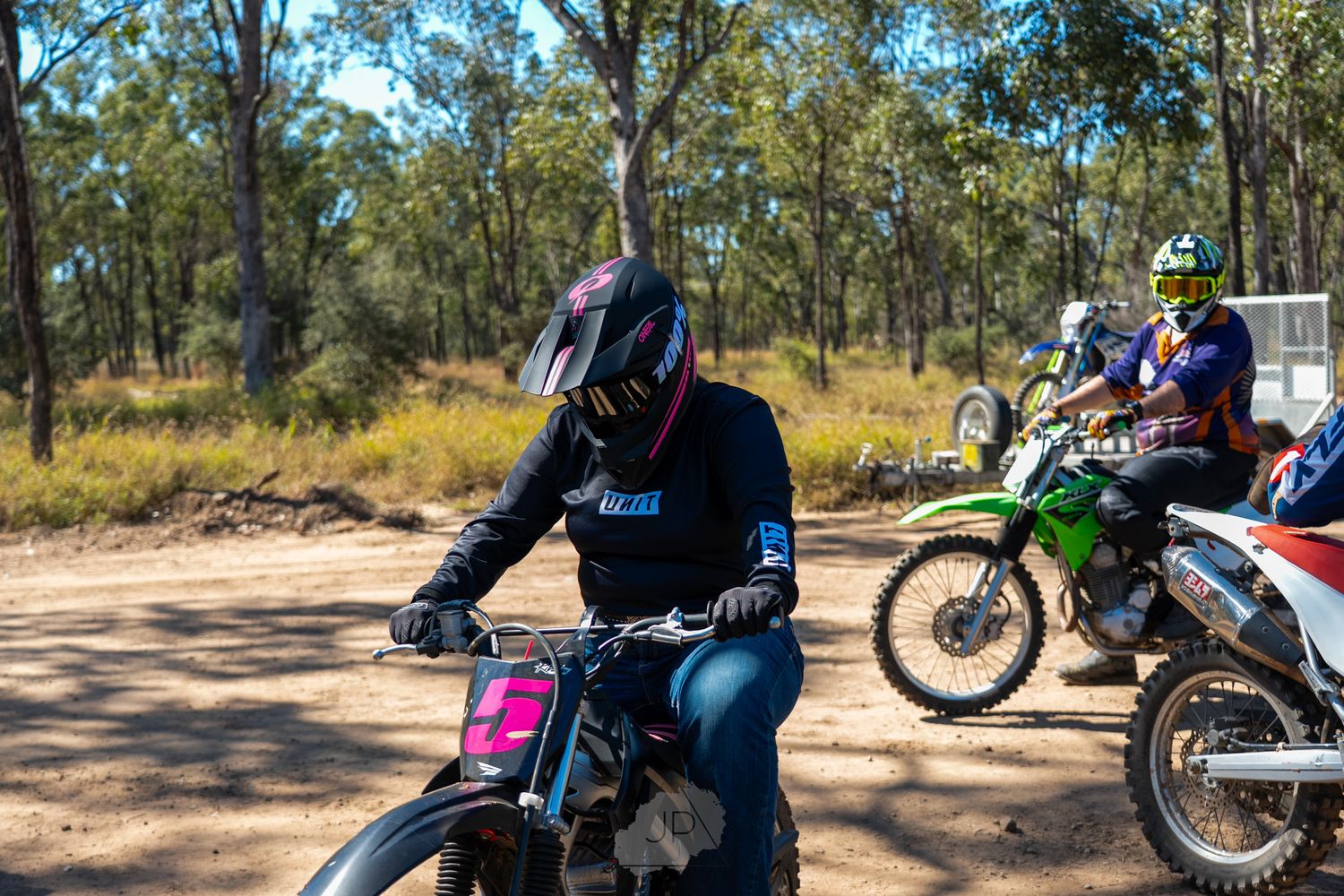 Dirt bike riders gathered on a trail in the Australian bush with motorcycles parked on dusty ground.