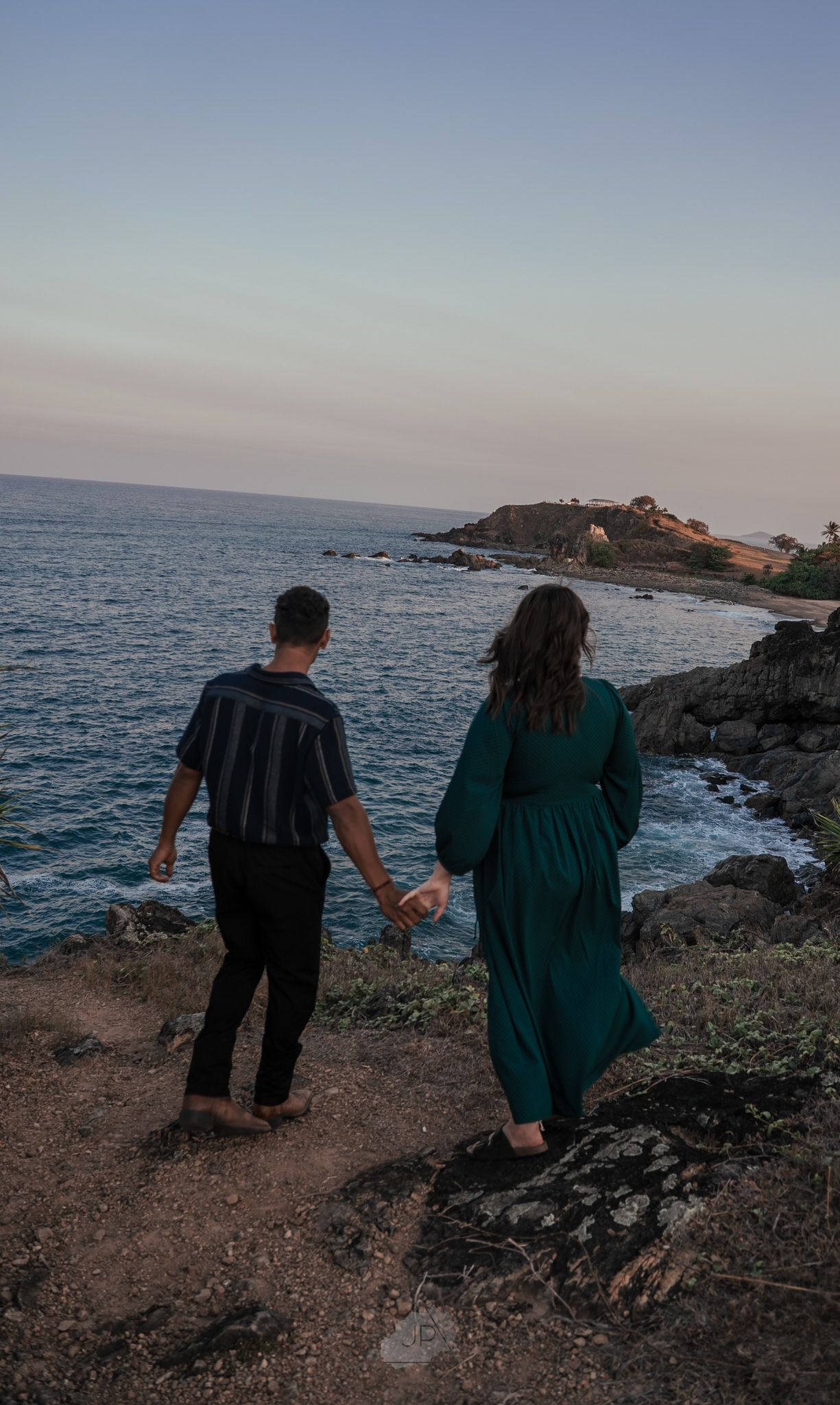 A couple walks along a rocky coastal path at sunset overlooking the ocean.