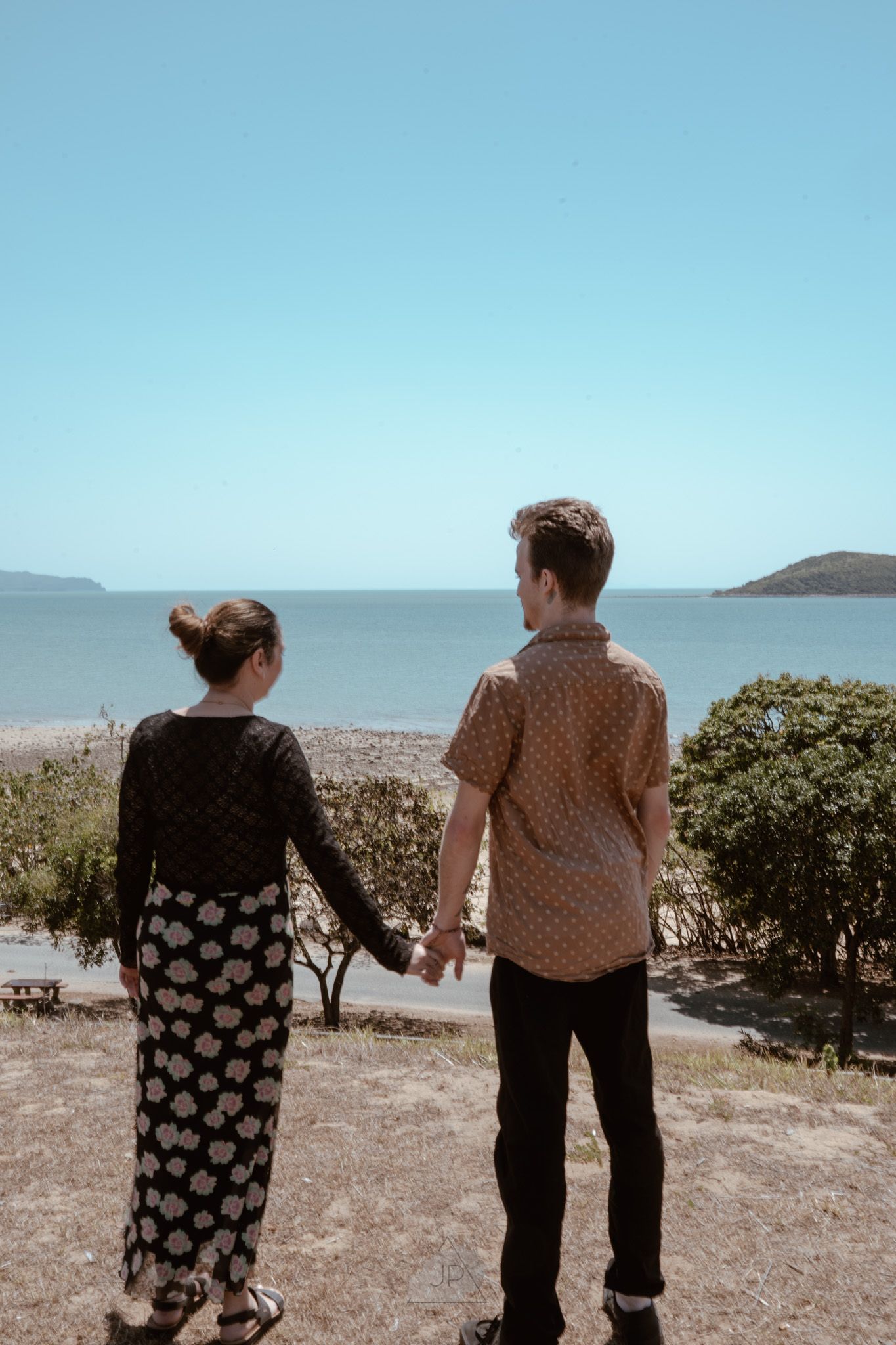 A couple holds hands while looking out at a tropical coastline with mountains in the distance.