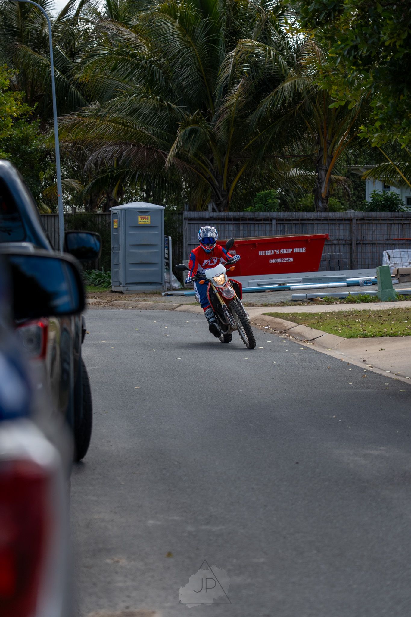 A motorcyclist rides on a street with palm trees and a portable toilet in the background.