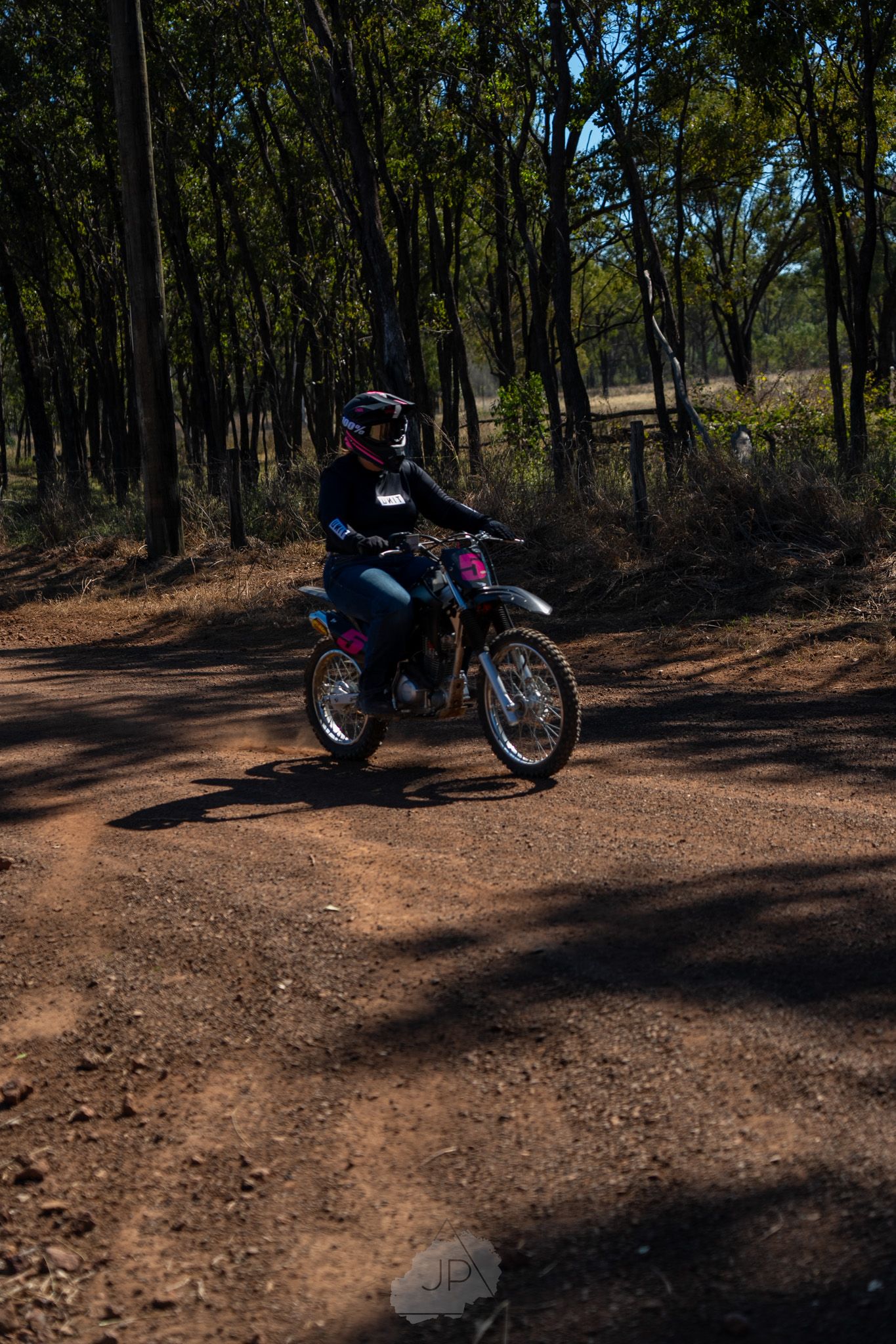 A dirt bike rider navigates through a wooded trail on a sunny day, casting shadows on the brown earth path.