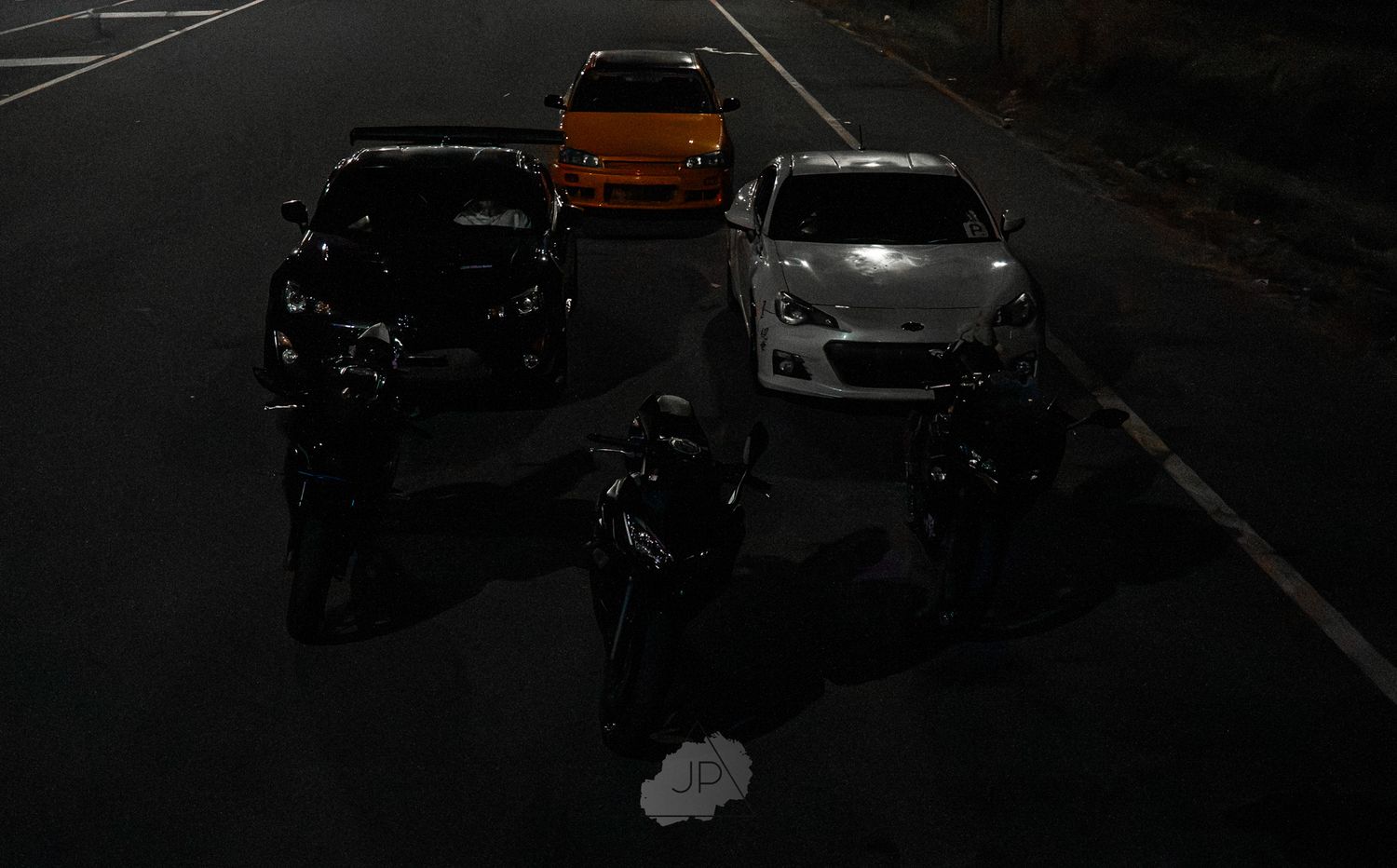 A motorcyclist stands between parked cars on a dark road at night with an amber light visible in the background.