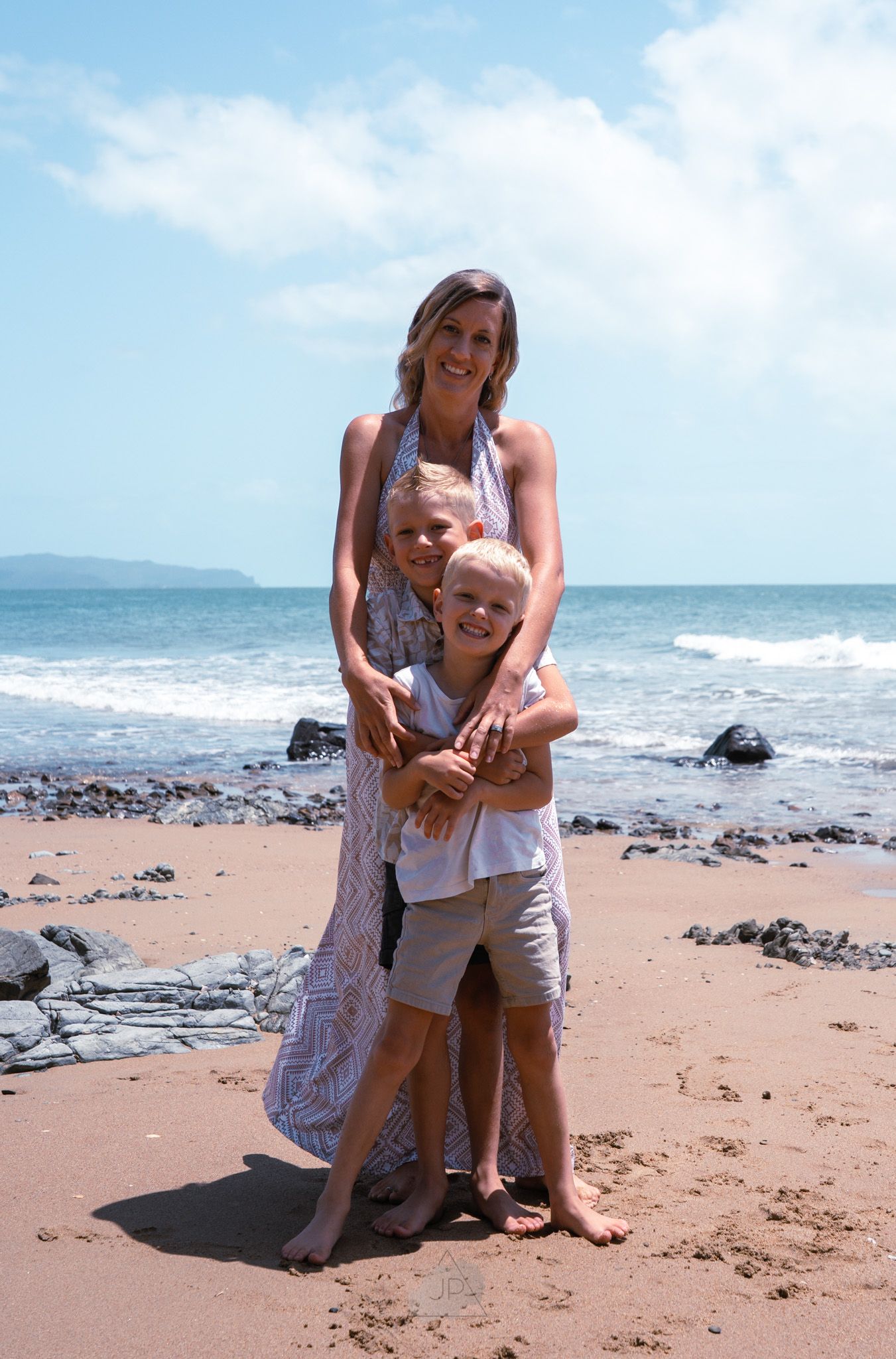 A family embraces while standing on a rocky beach with blue ocean waves and mountains in the background.