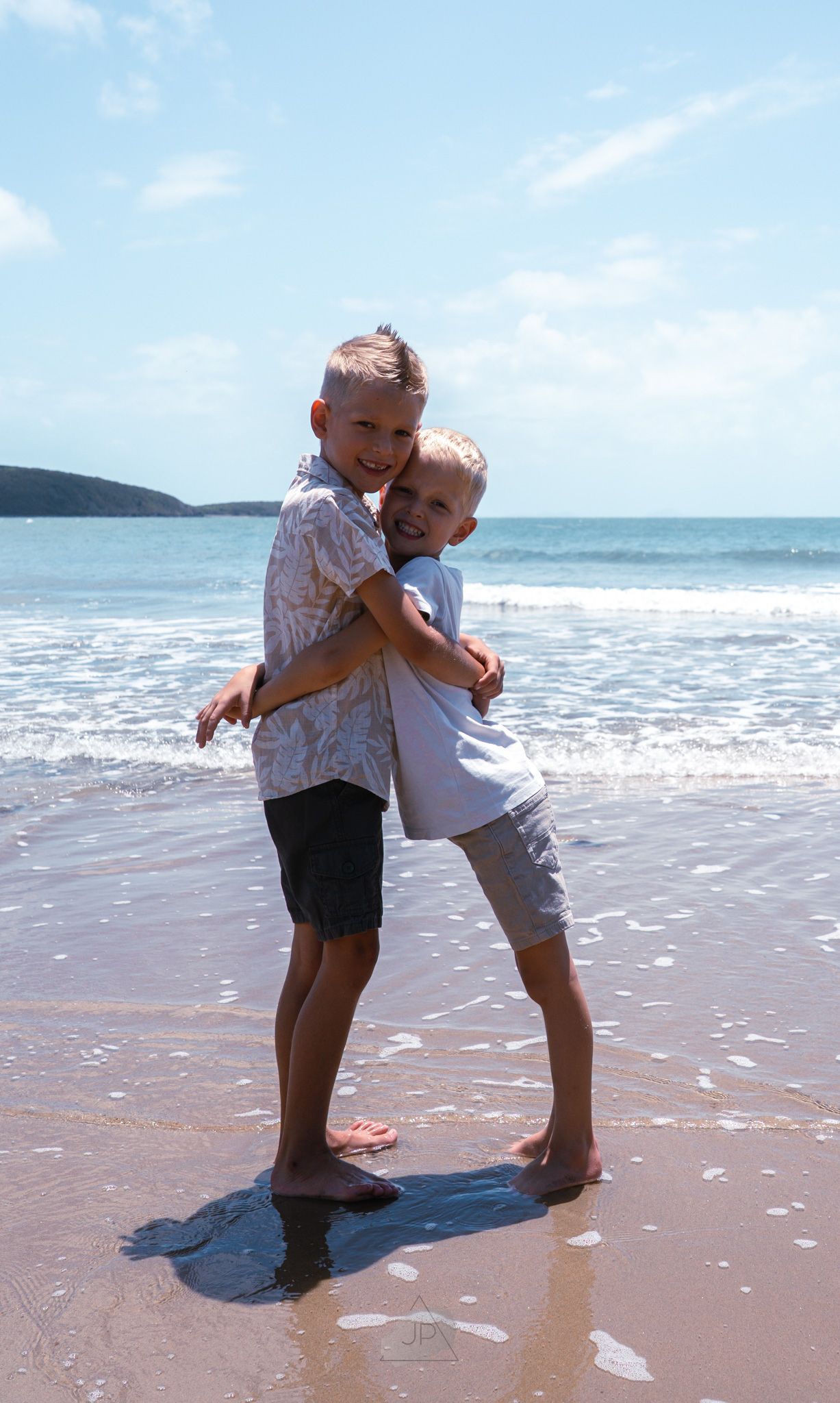 Two children play and hug on the wet sand at the beach with gentle waves rolling in behind them.