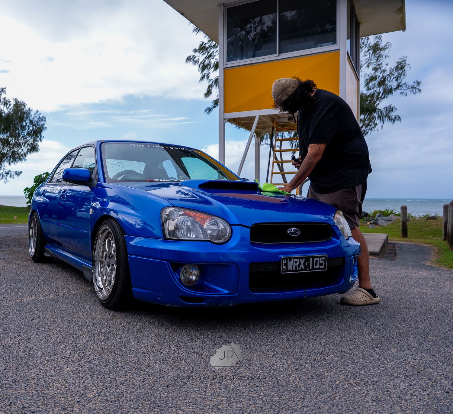 Blue modified Subaru WRX sitting low on aftermarket wheels parked at a gas station with scenic coastal view.