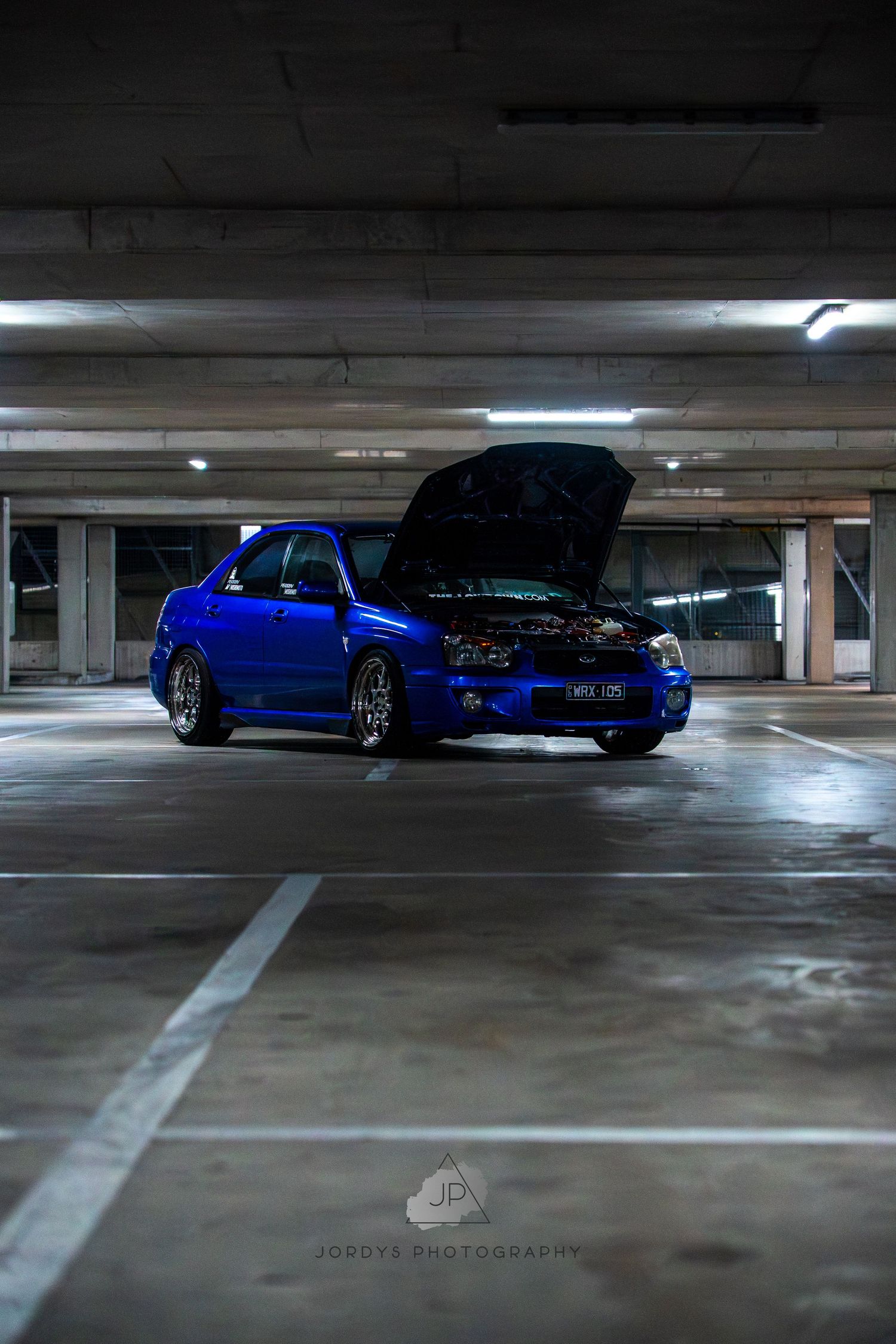 Dark parking garage showcasing a blue sports car parked in dramatic low lighting with luxury car spotlight reflections.