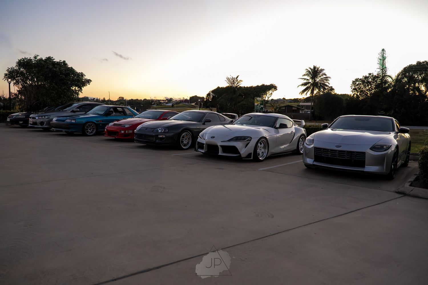 Rows of sleek luxury sports cars parked at sunset in a dealer lot with palm trees in the background.