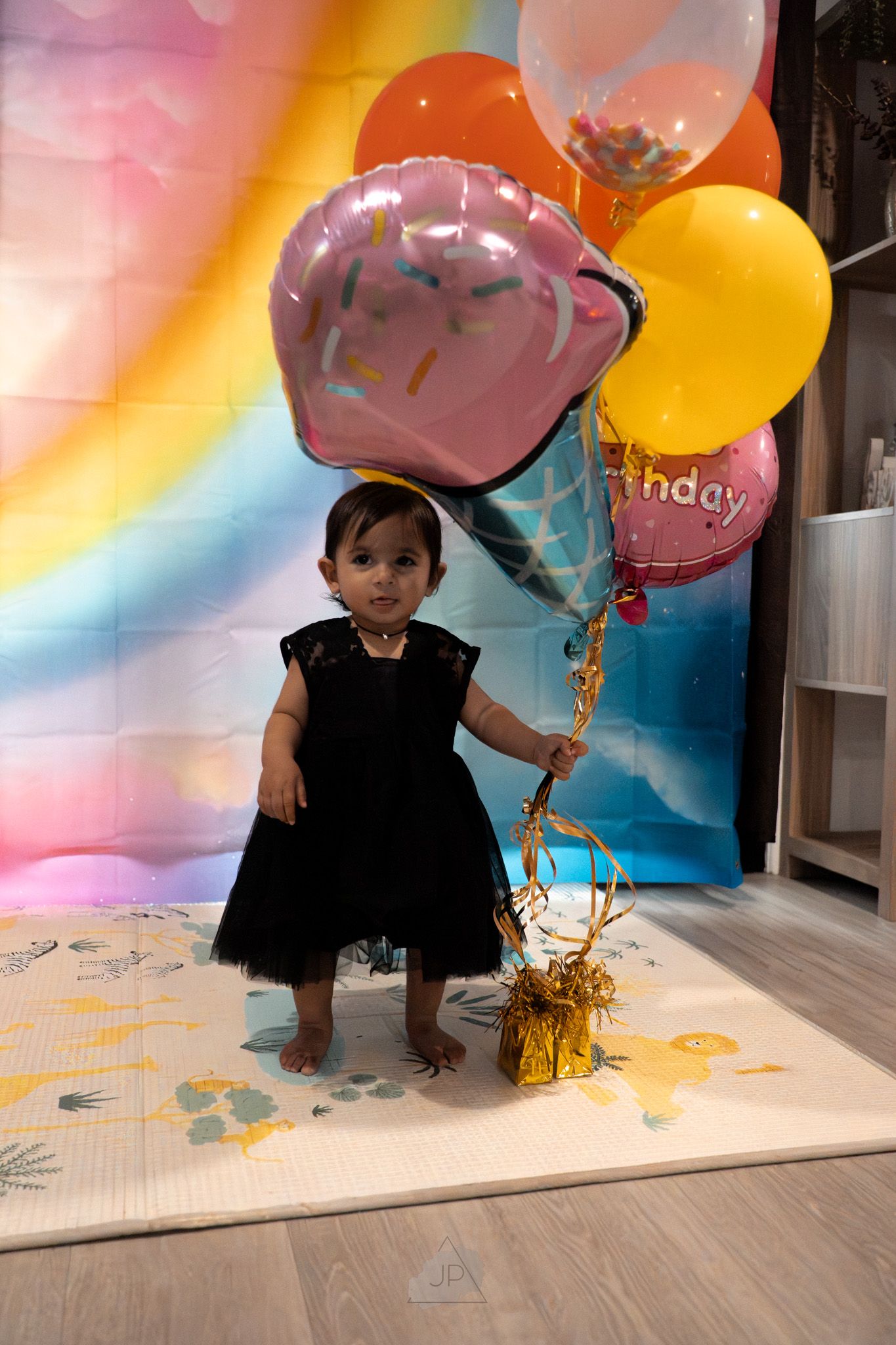 Pink and yellow balloons float against a colorful backdrop as a small child plays on a white floor.