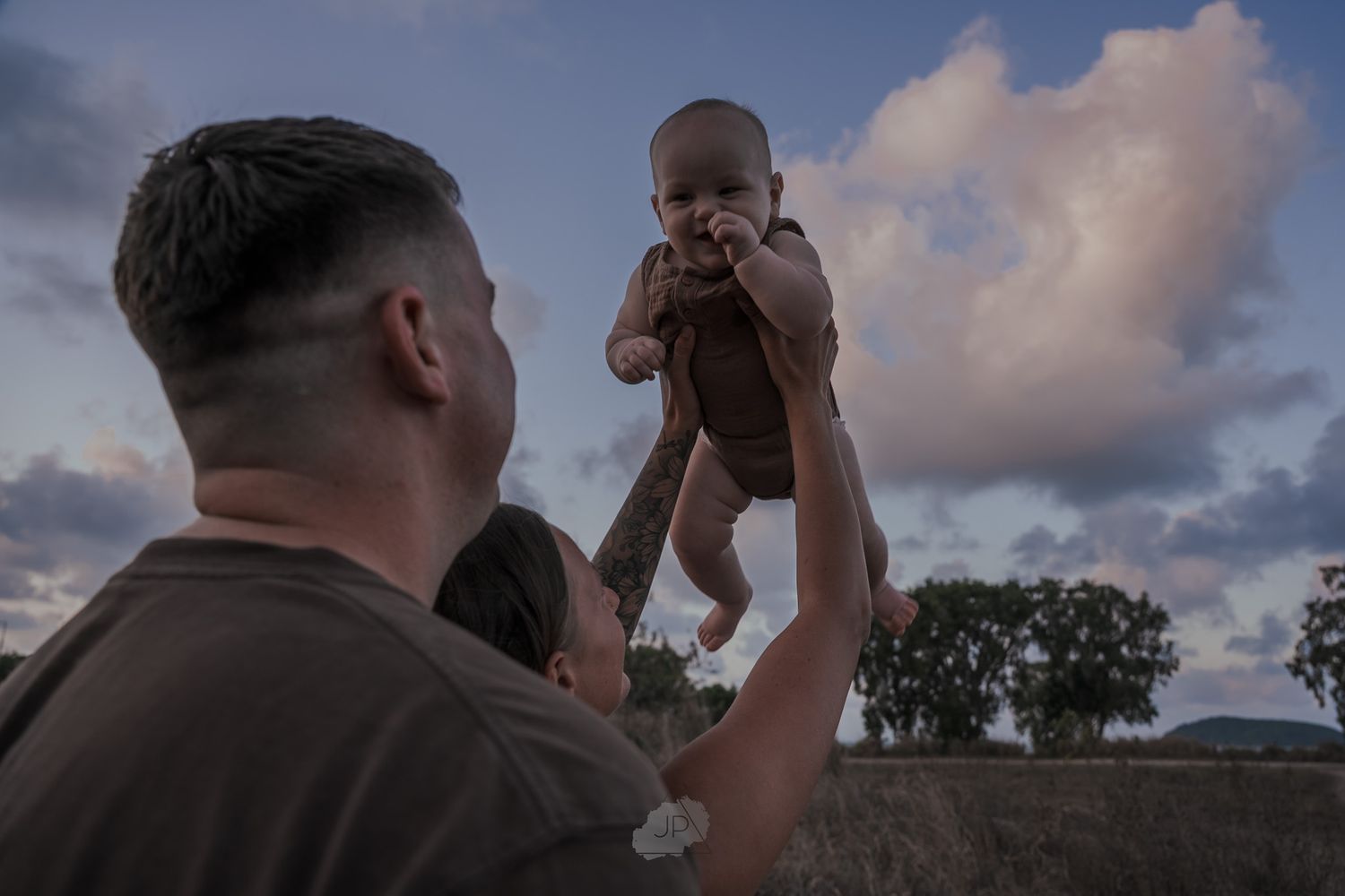 A person lifts a baby up against a dramatic cloudy sunset sky with silhouetted trees in the background.