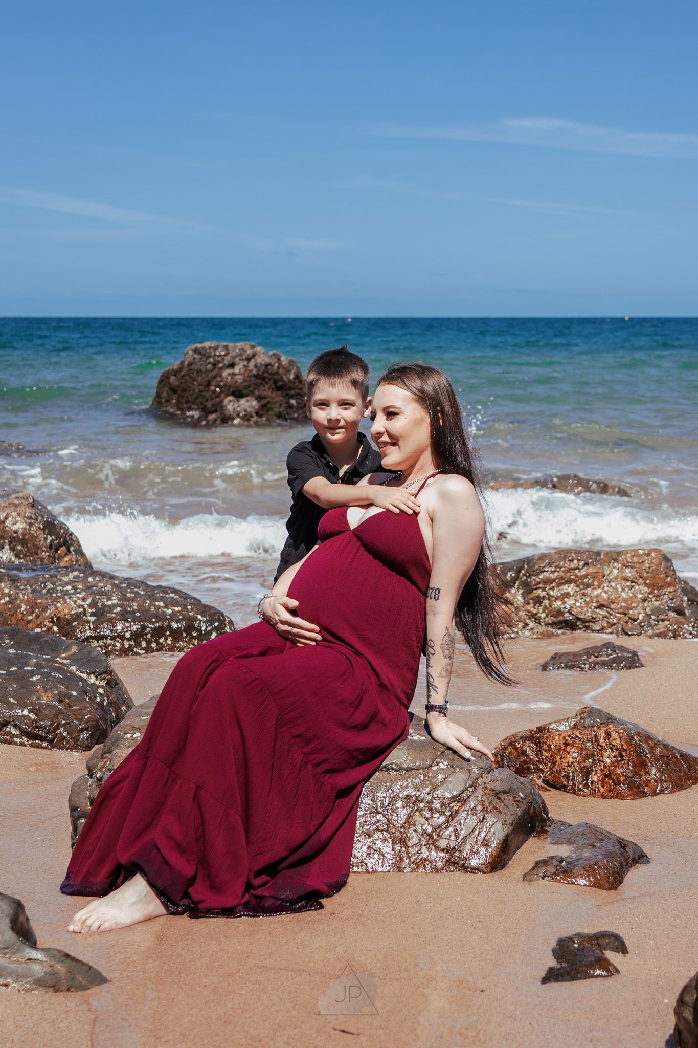 A couple in burgundy attire poses romantically on a sandy beach with waves and rocks in the background.