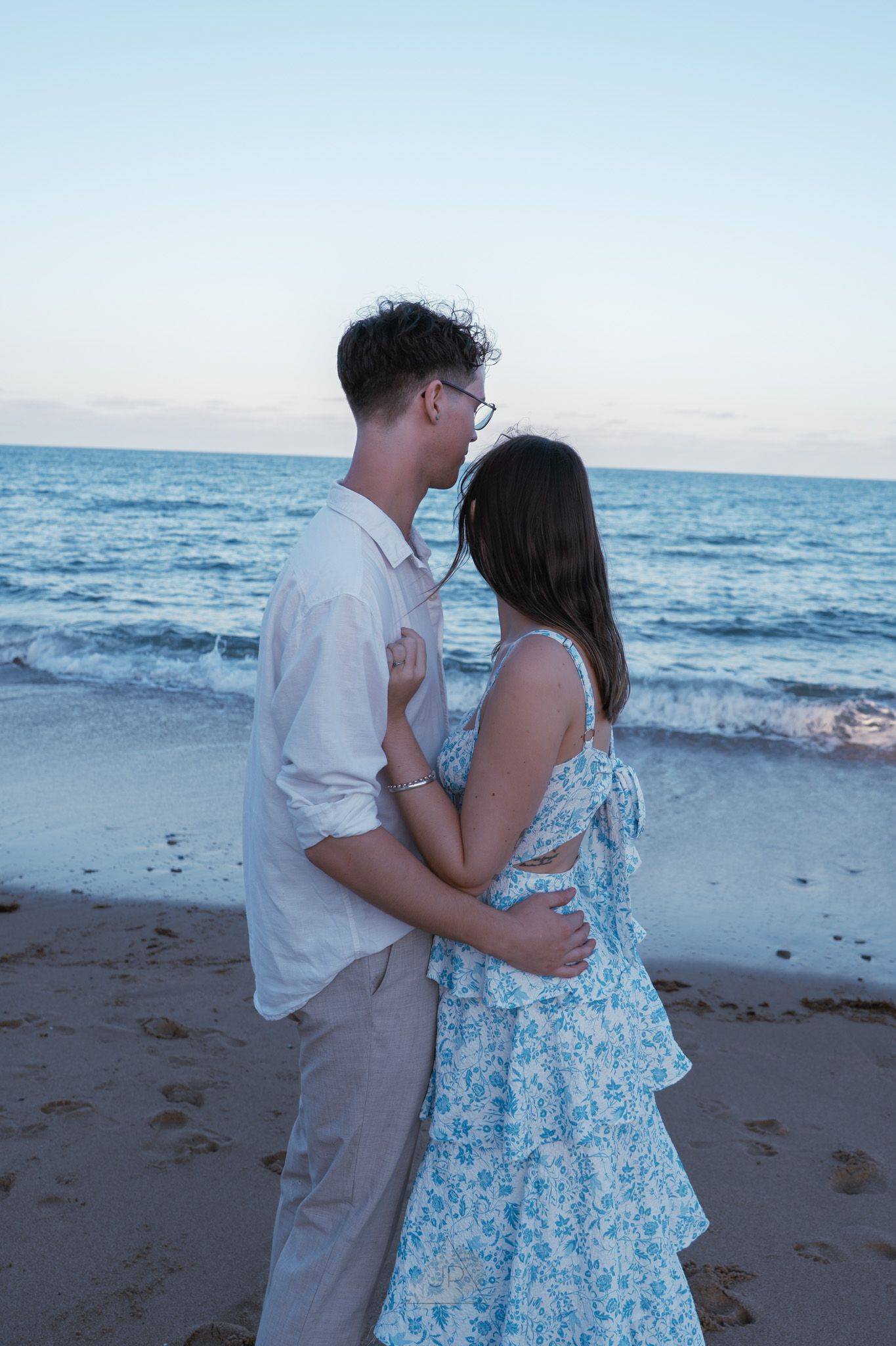 A couple shares a romantic moment on the beach at sunset with waves crashing in the background.