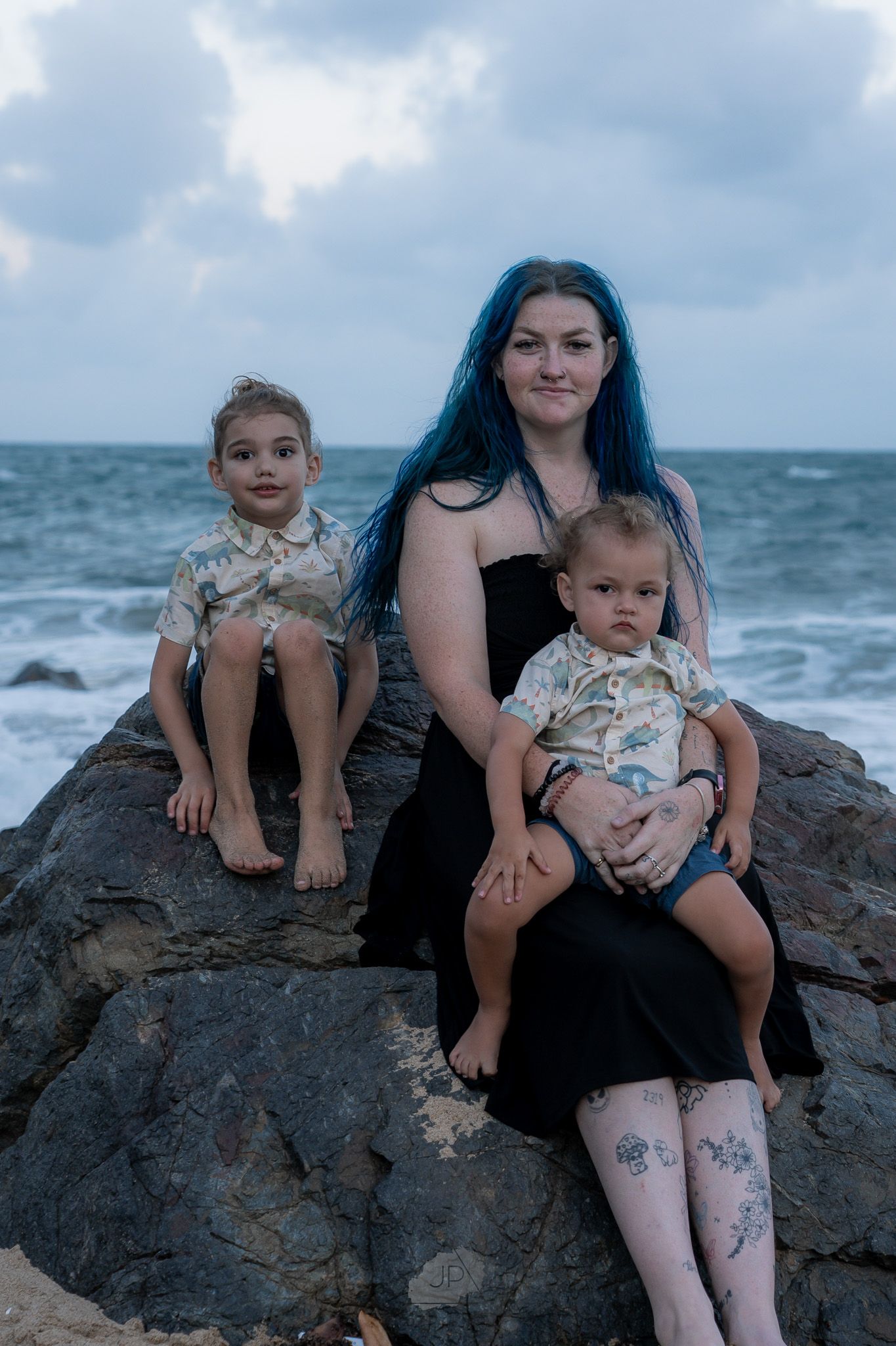 Family sitting together on coastal rocks at the beach with stormy ocean waves in the background.