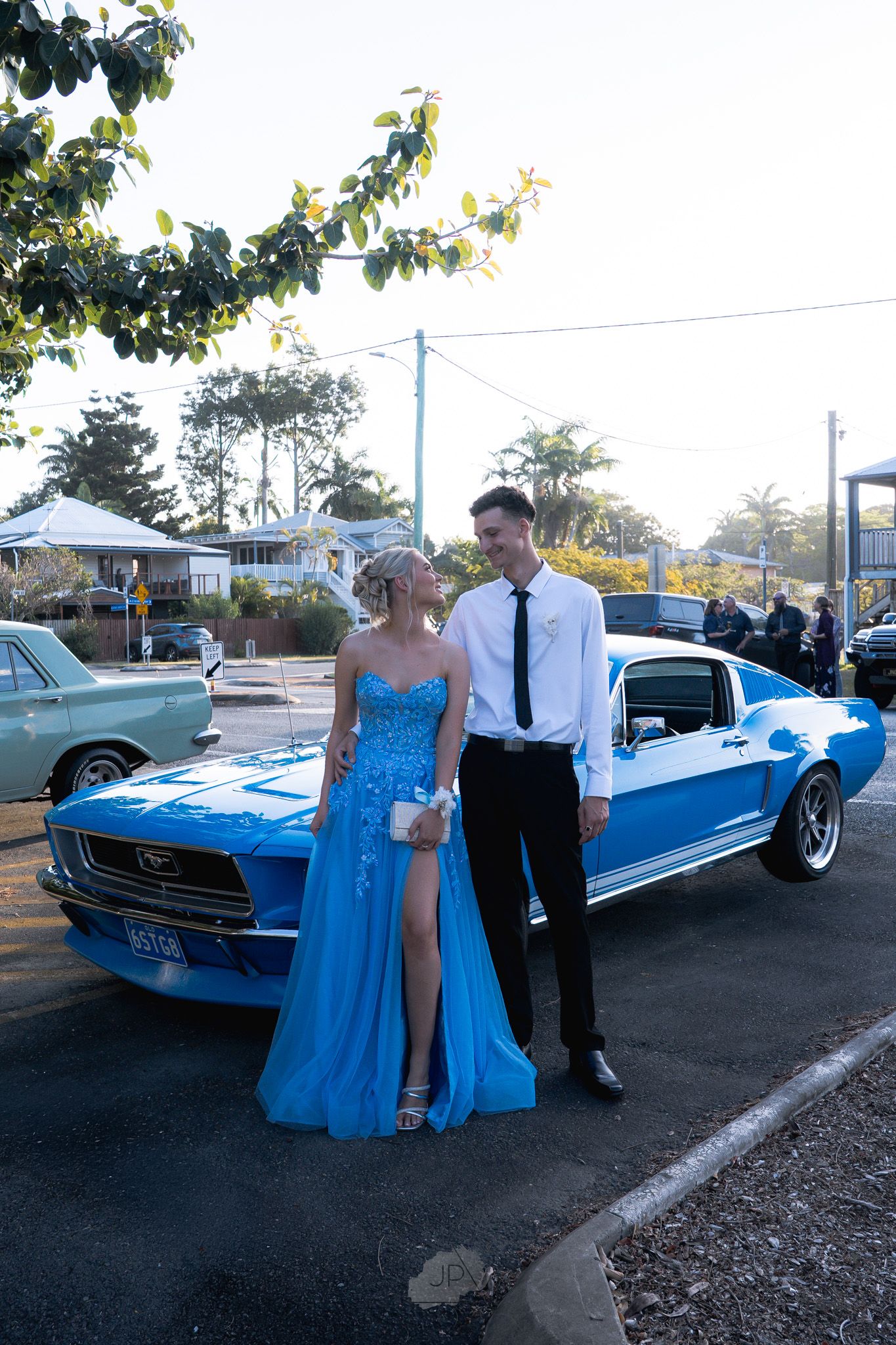 A couple poses next to a vintage blue Ford Mustang at sunset in formal attire.