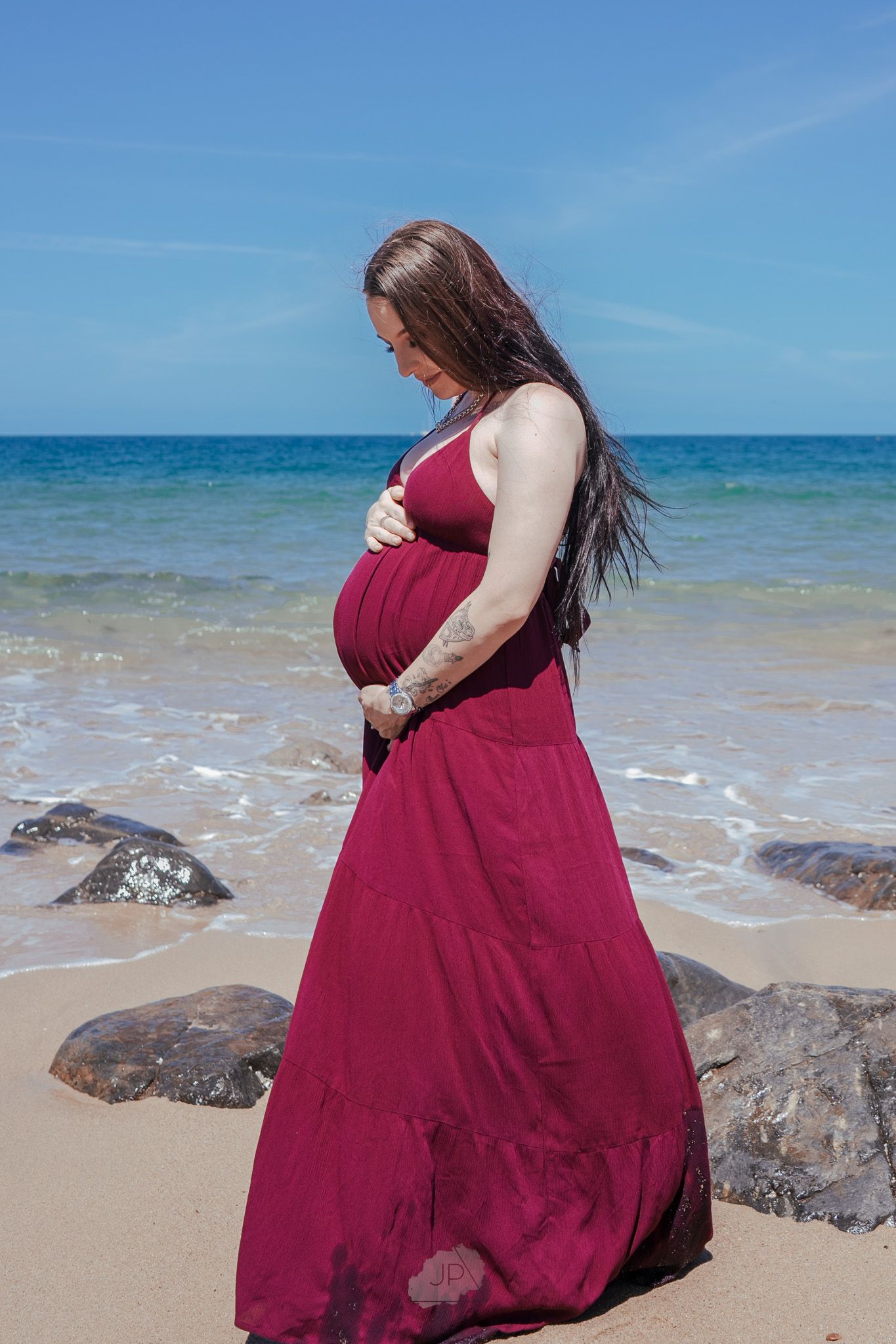 A woman in a burgundy dress poses on a rocky beach with turquoise ocean waters and clear blue skies in the background.
