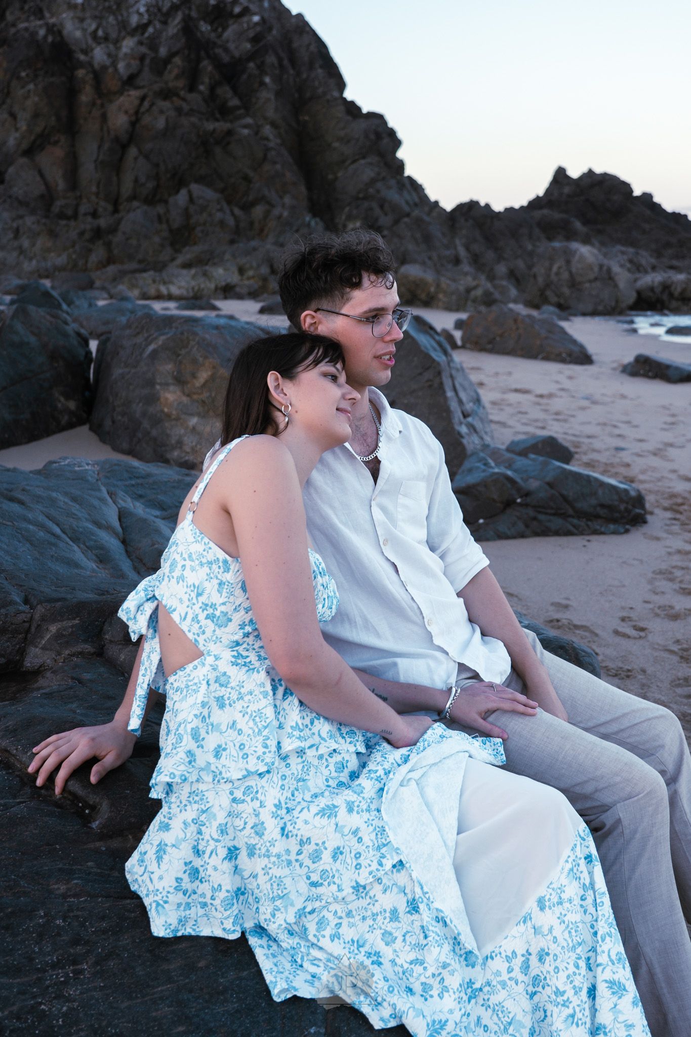 A couple in blue and white attire sits together on coastal rocks at sunset sharing intimate moments on the beach.