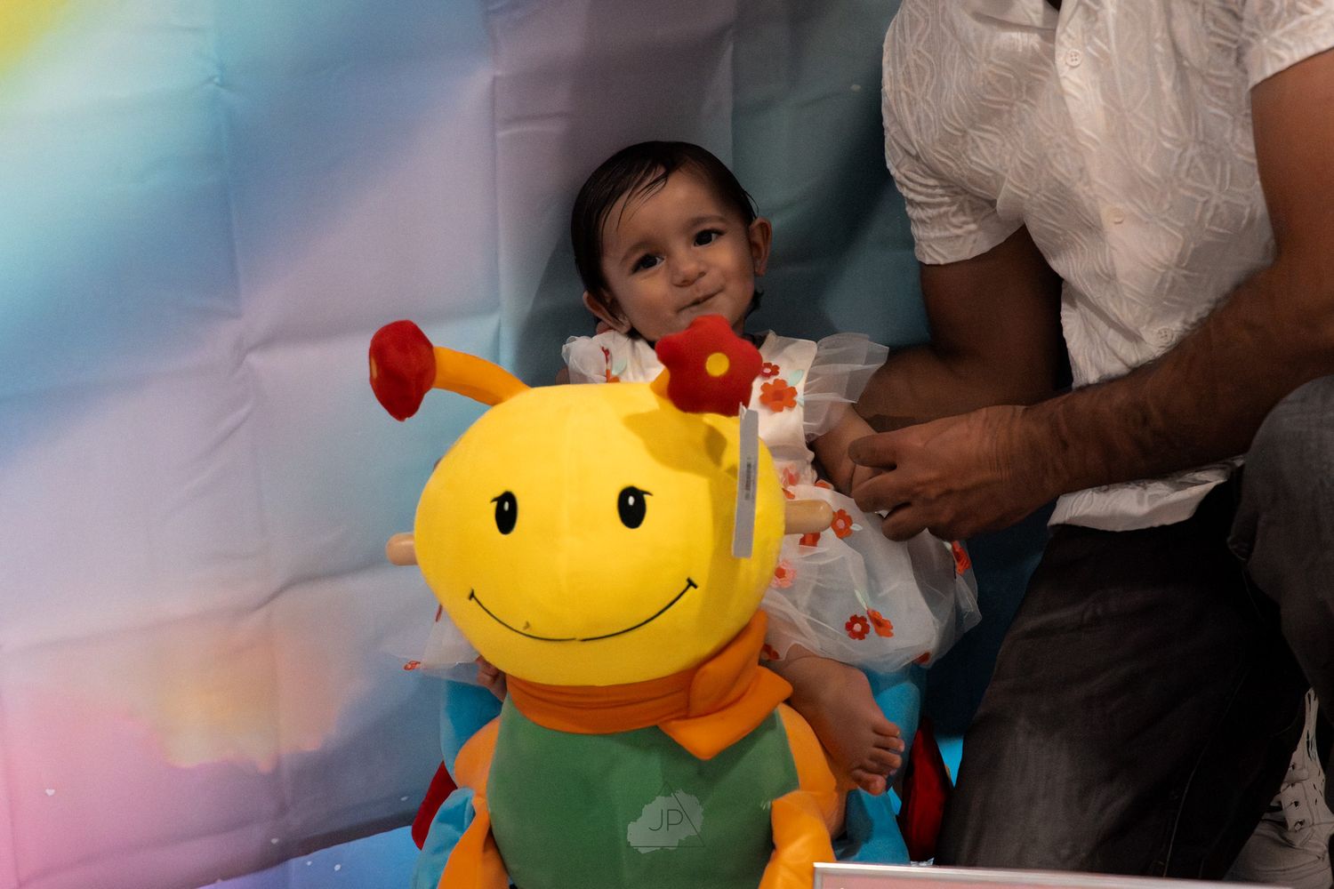 A colorful plush smiley face toy with antennae sits next to someone in a bright rainbow-colored setting.