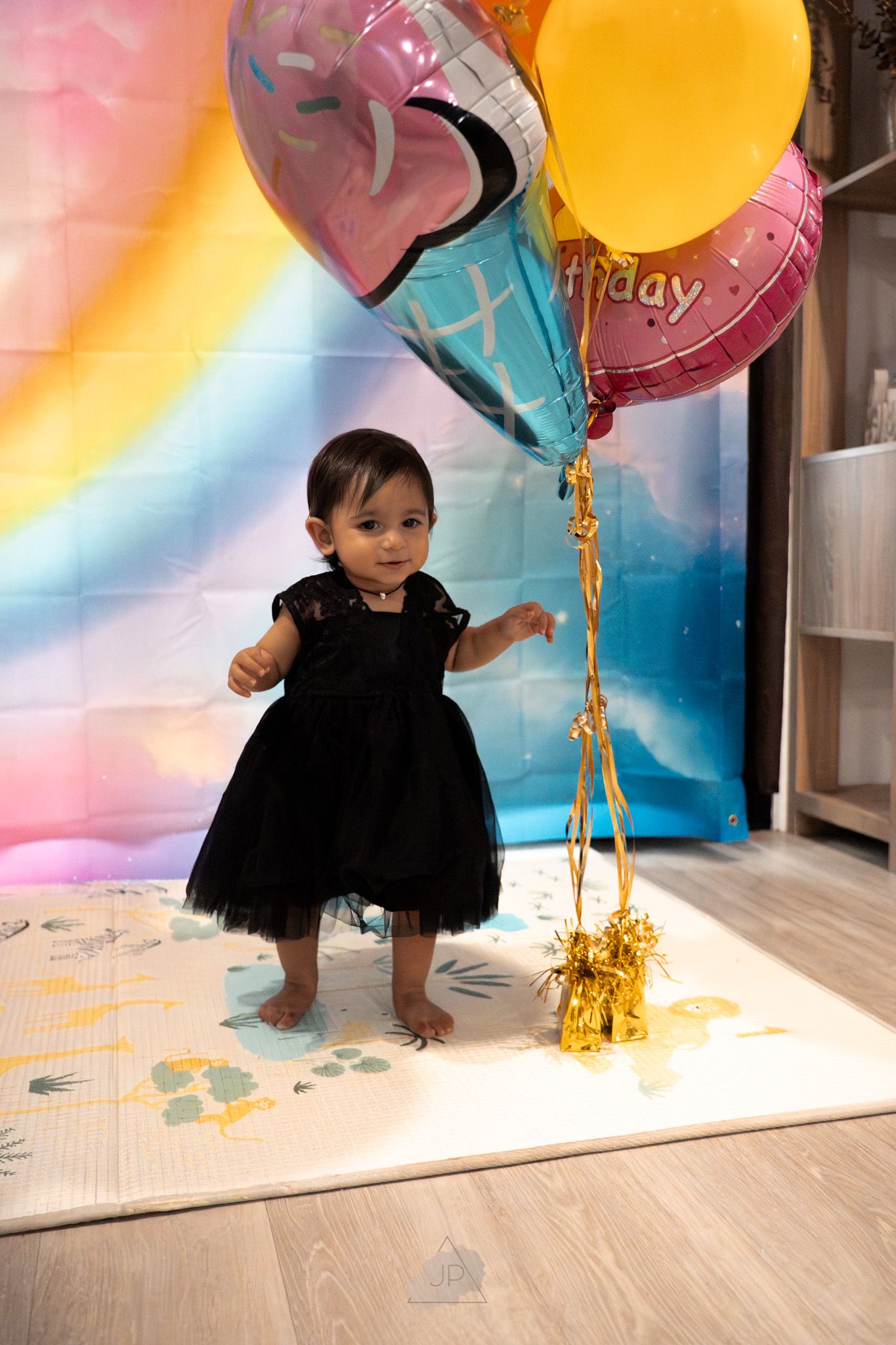 A small child in a black tutu dress crawls under colorful balloons during a birthday celebration with rainbow backdrop.