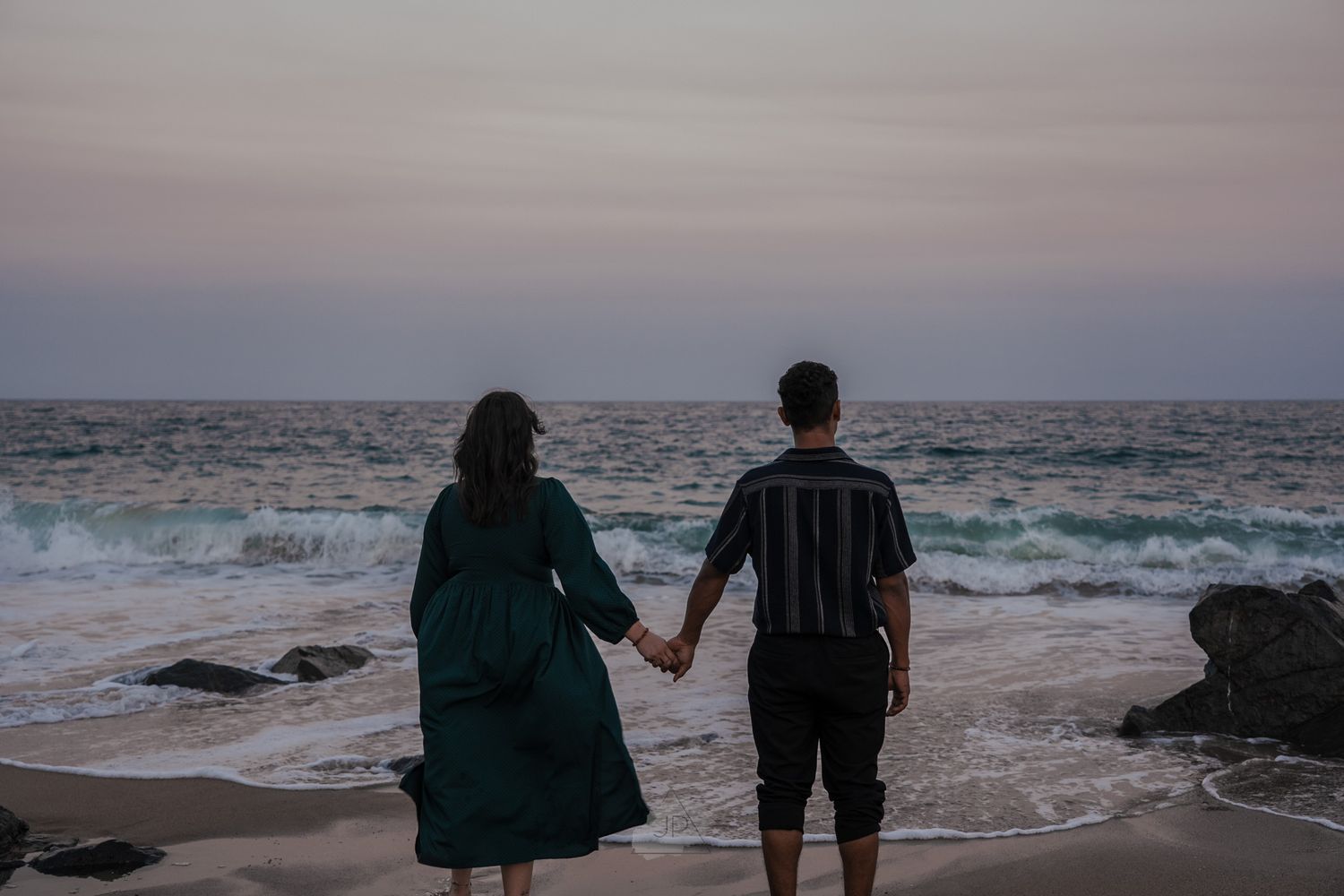 A couple holds hands while watching the sunset over crashing ocean waves from a rocky beach.