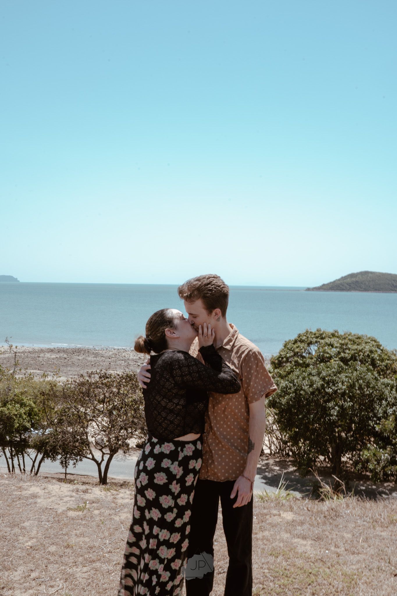 A couple shares a tender moment on a hilltop overlooking the ocean on a sunny day.