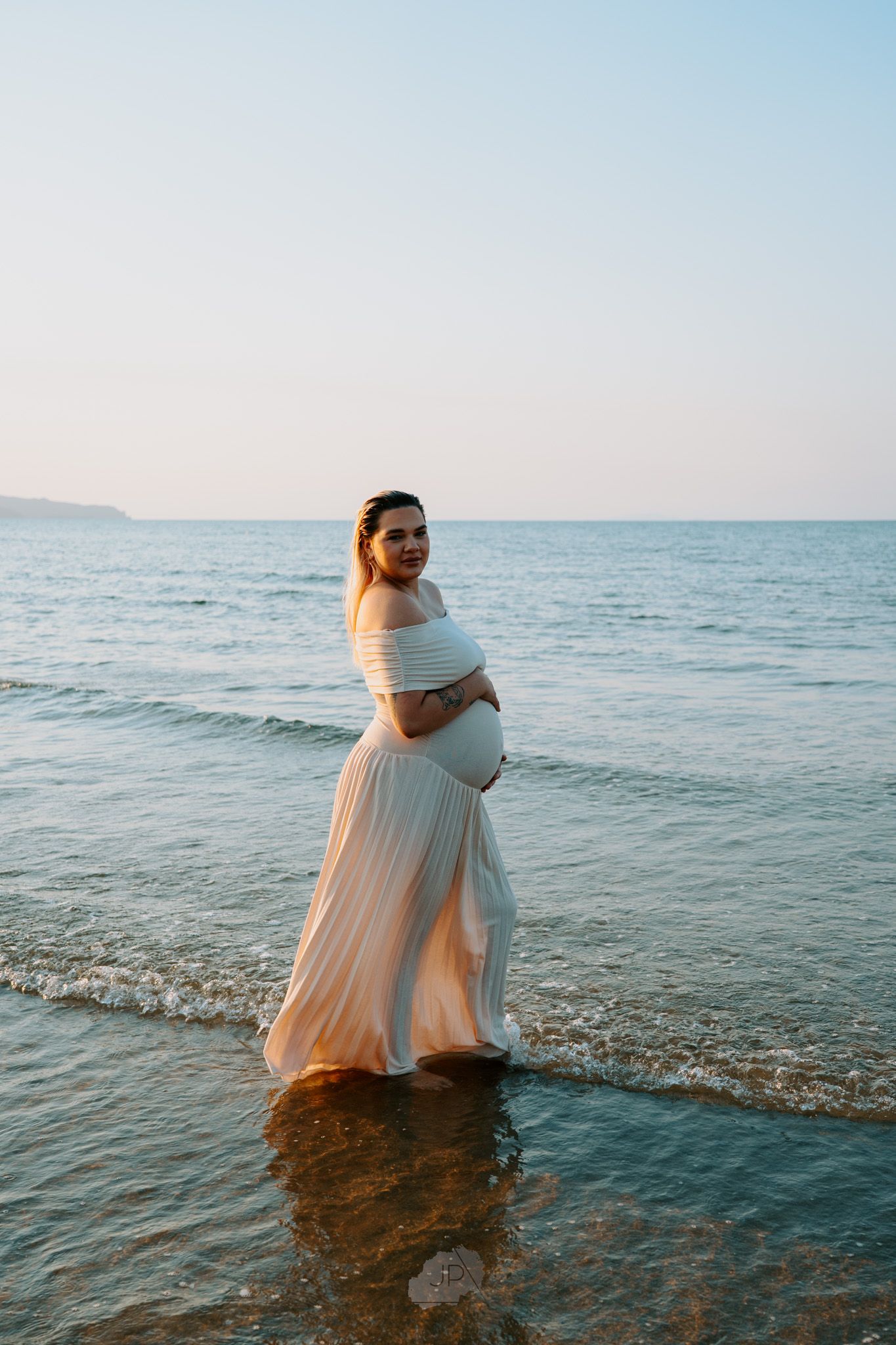 A person in a flowing white dress stands in shallow ocean water at sunset creating a dreamy coastal scene.
