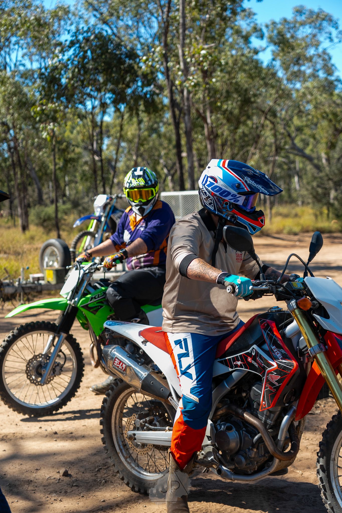Four dirt bikes parked in a row on dusty terrain with trees and bushes in the background on a sunny day.