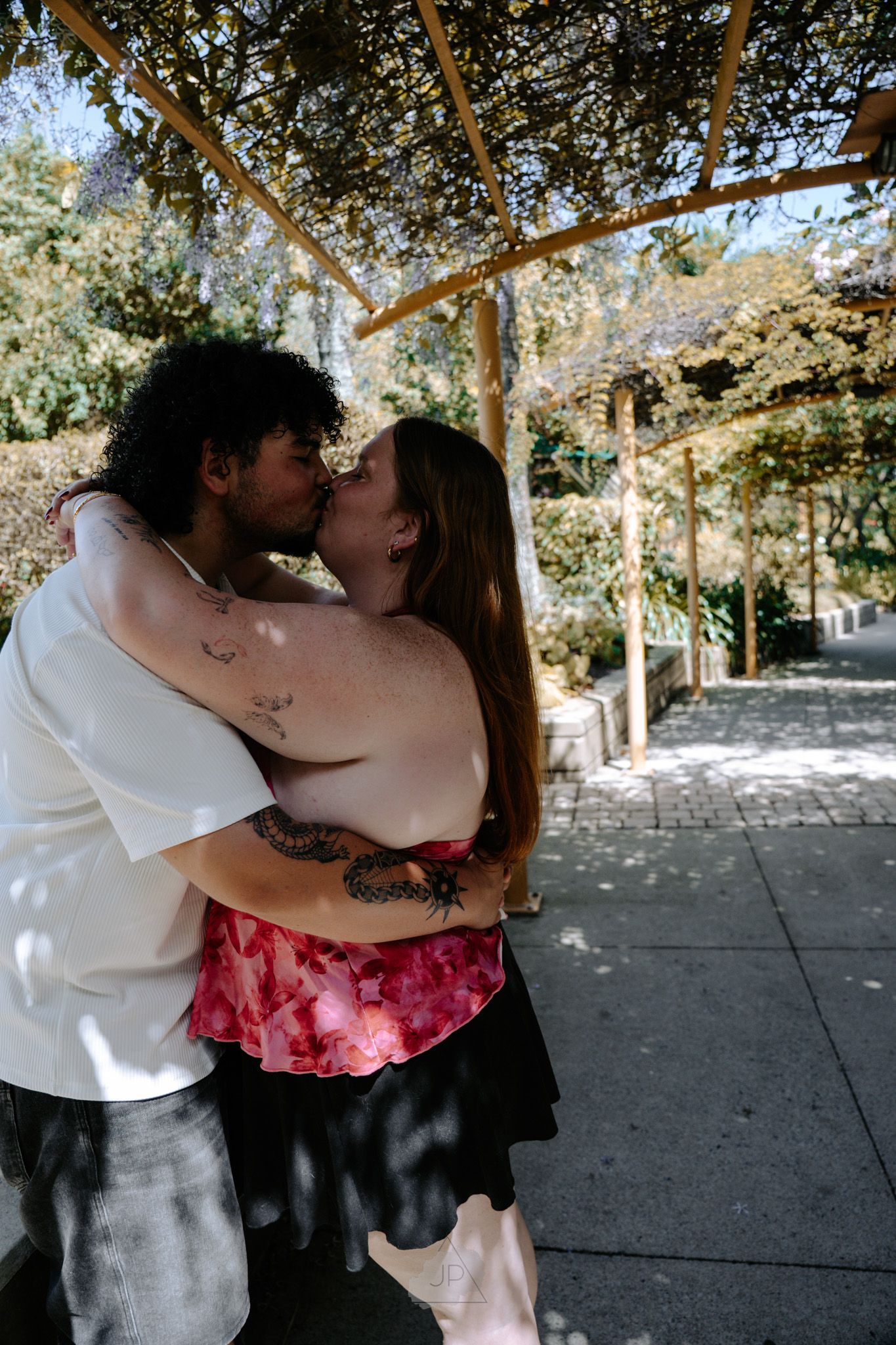 A romantic couple shares intimate kisses under a wooden pergola in an outdoor setting with dappled sunlight.