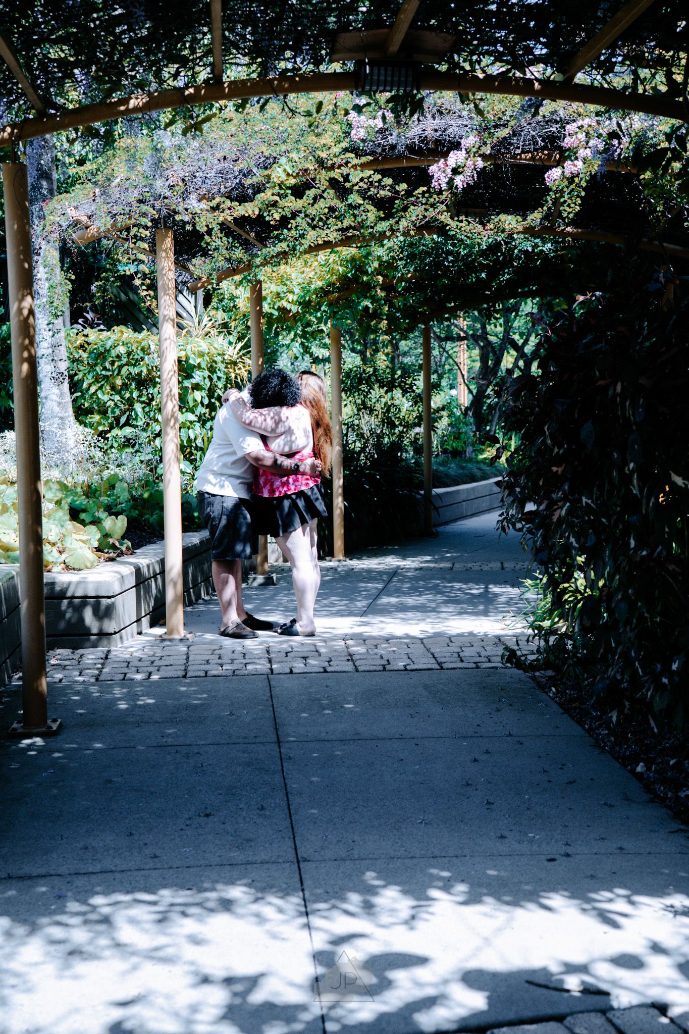 A long covered walkway with stone pavers winds through a lush garden setting on a sunny day.