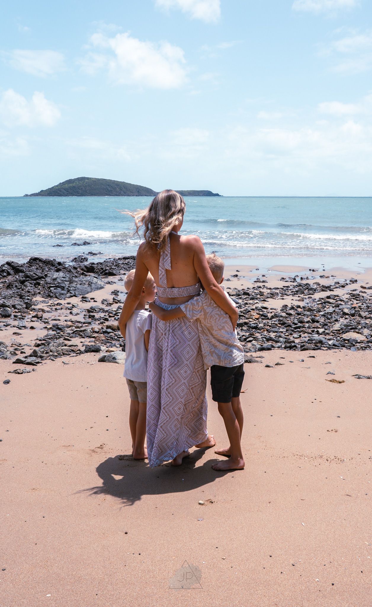 People sitting on a sandy beach with a rocky shoreline and island visible in the distance on a sunny day.