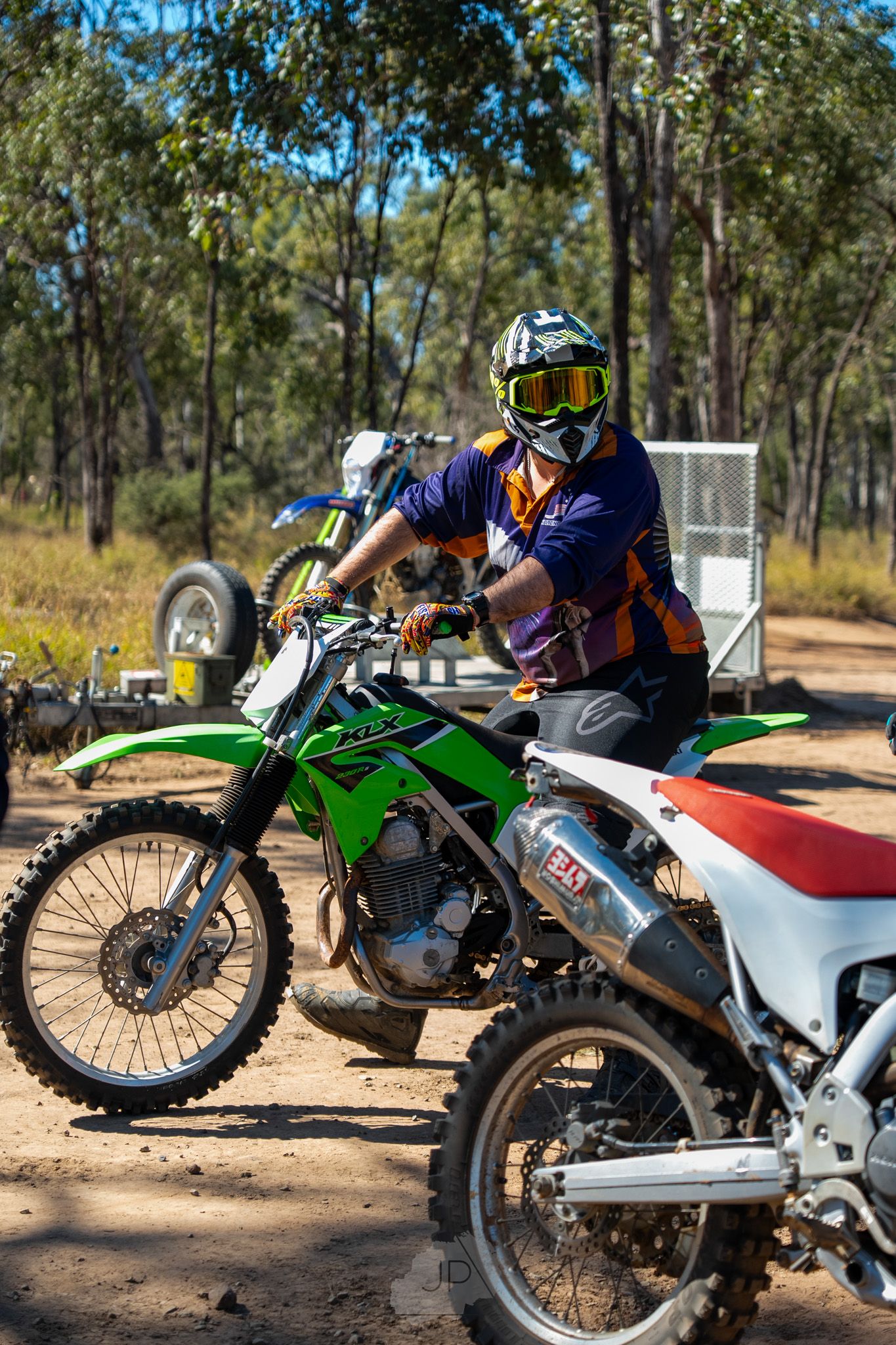 Kawasaki dirt bikes parked in a line at a wooded trail area with riders preparing for off-road adventure.