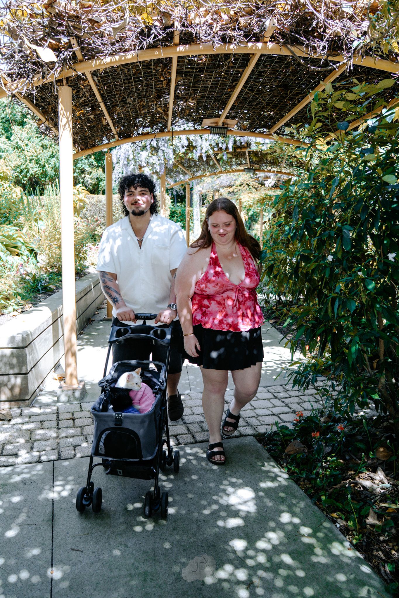 Two people sitting together under a shaded pergola in a garden setting with stone pavers and greenery.