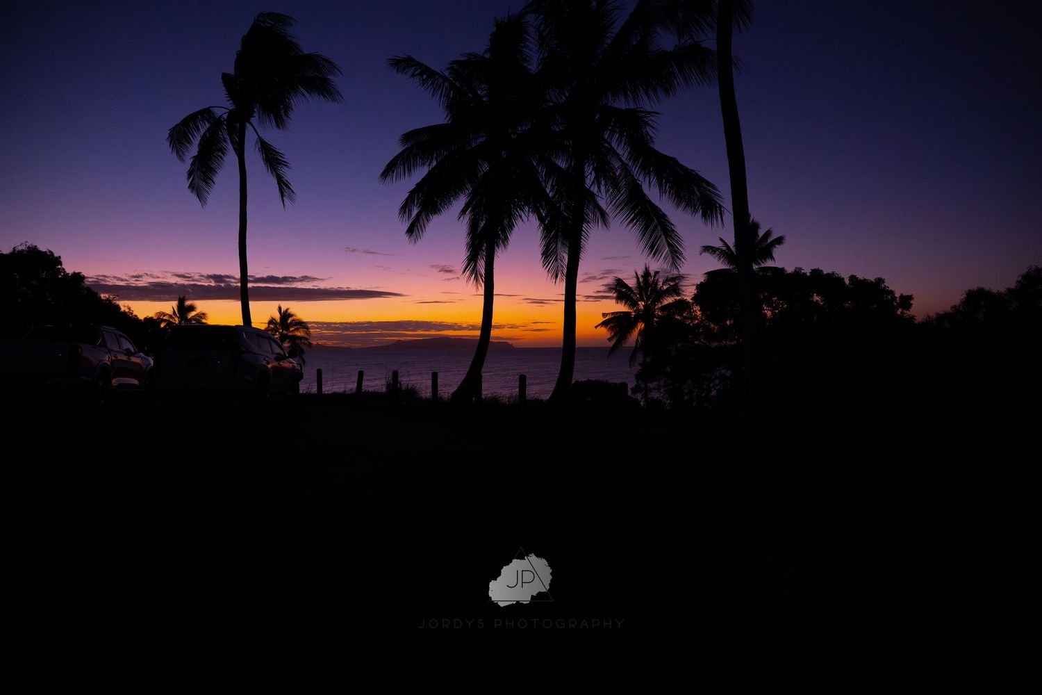 Silhouetted palm trees stand against a vibrant purple and orange sunset sky over a tropical beach.