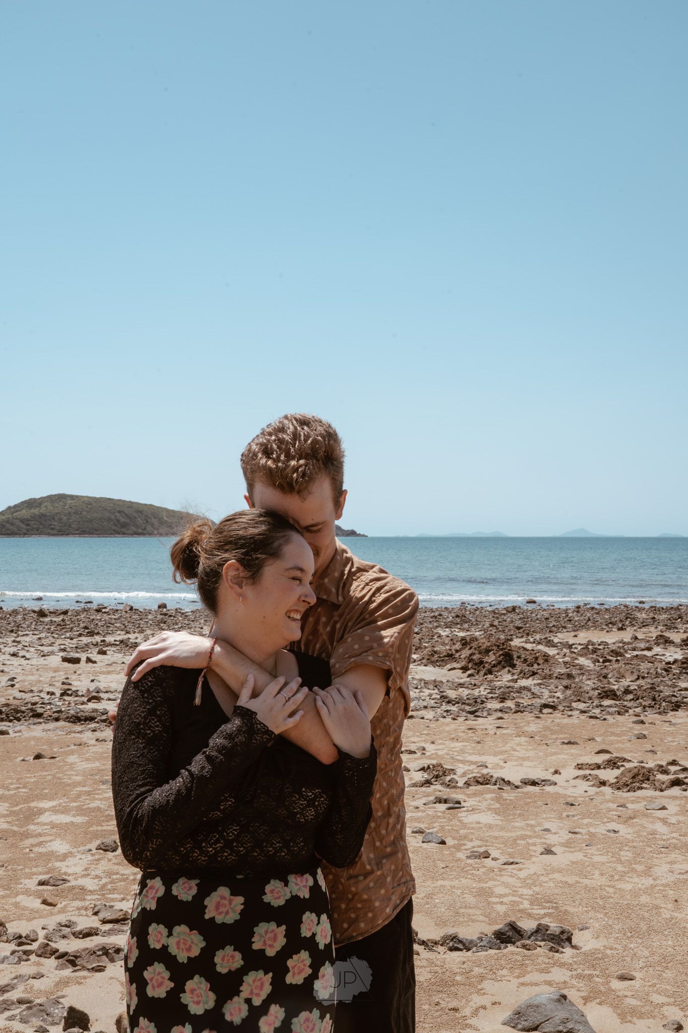 A couple embraces on a rocky beach shore with blue ocean and clear sky in the background.