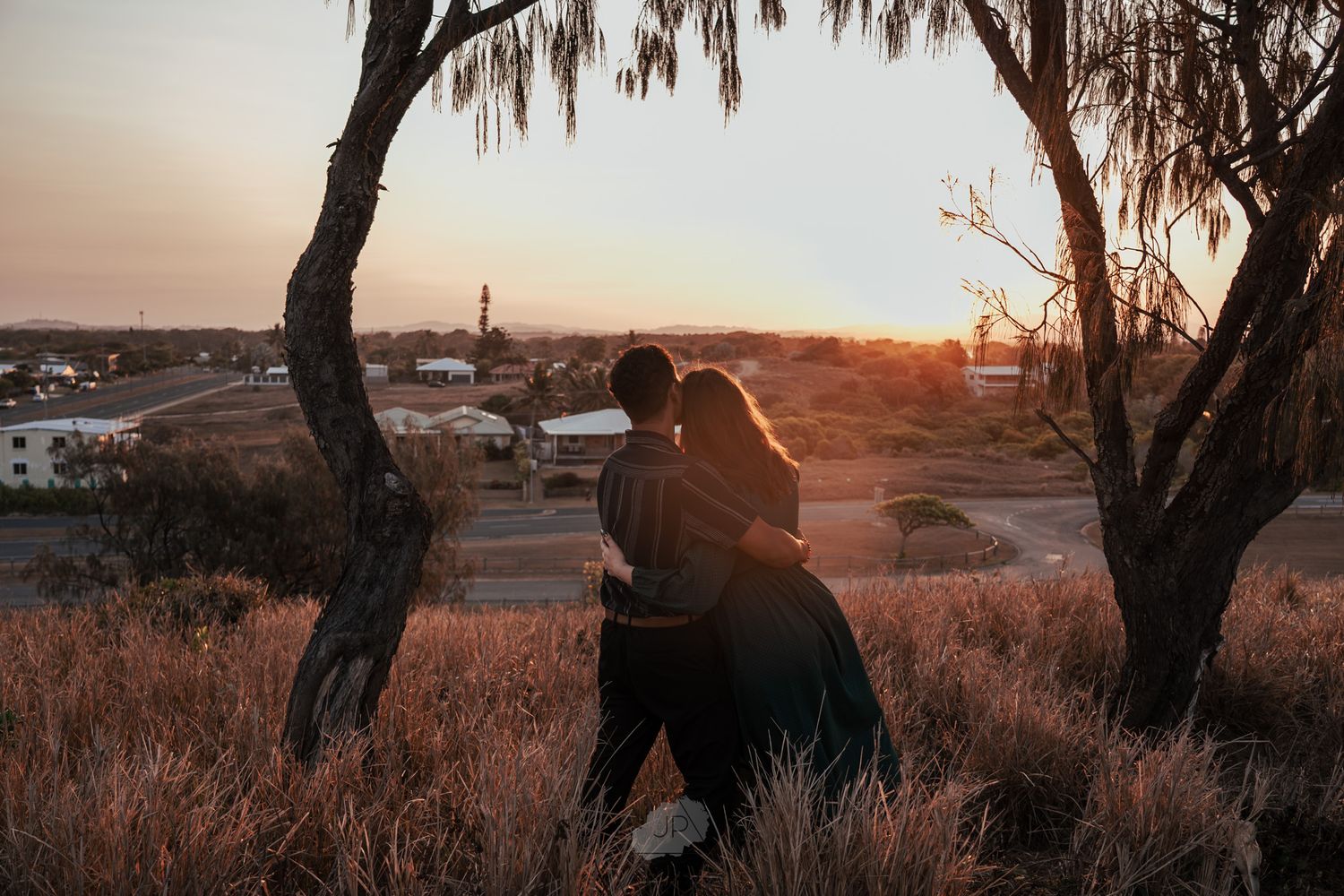 Silhouetted trees frame a golden sunset over a rural landscape with rolling hills and scattered buildings in the distance.