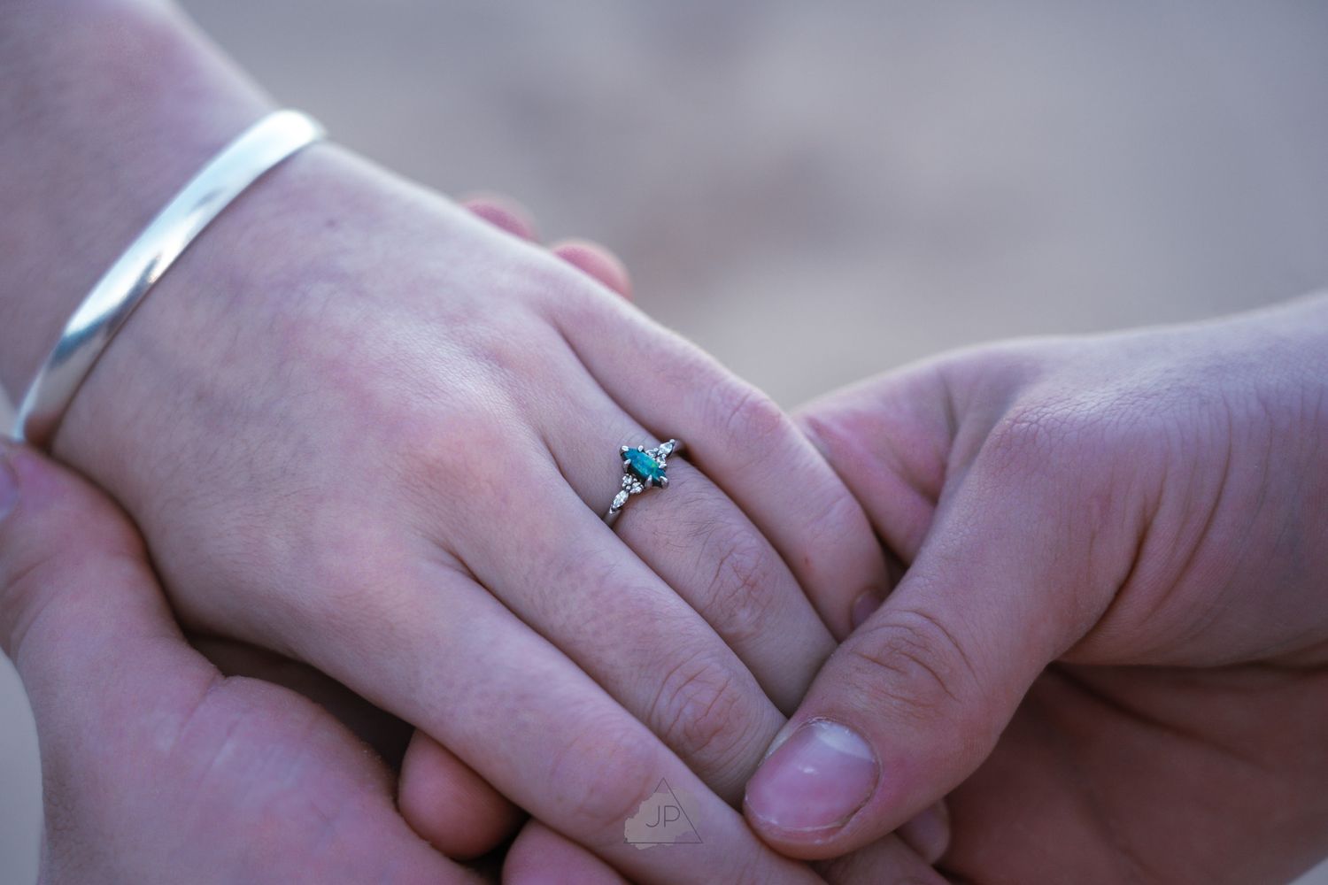 Couple's intertwined hands displaying a turquoise engagement ring against a soft blurred background.