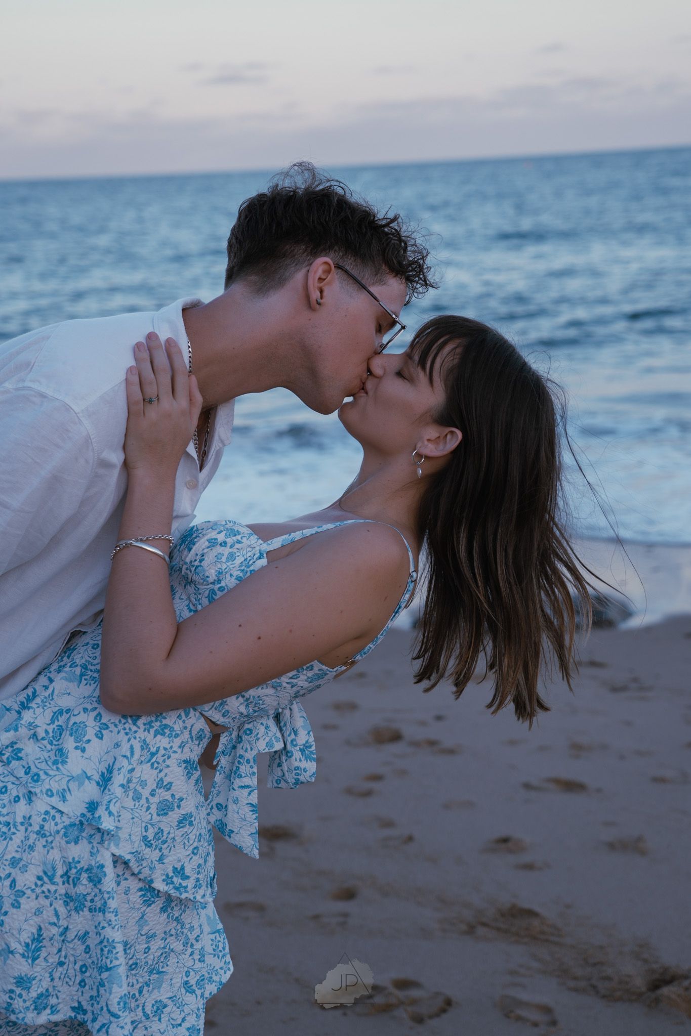A couple shares a tender moment on the beach at dusk, with ocean waves in the background.