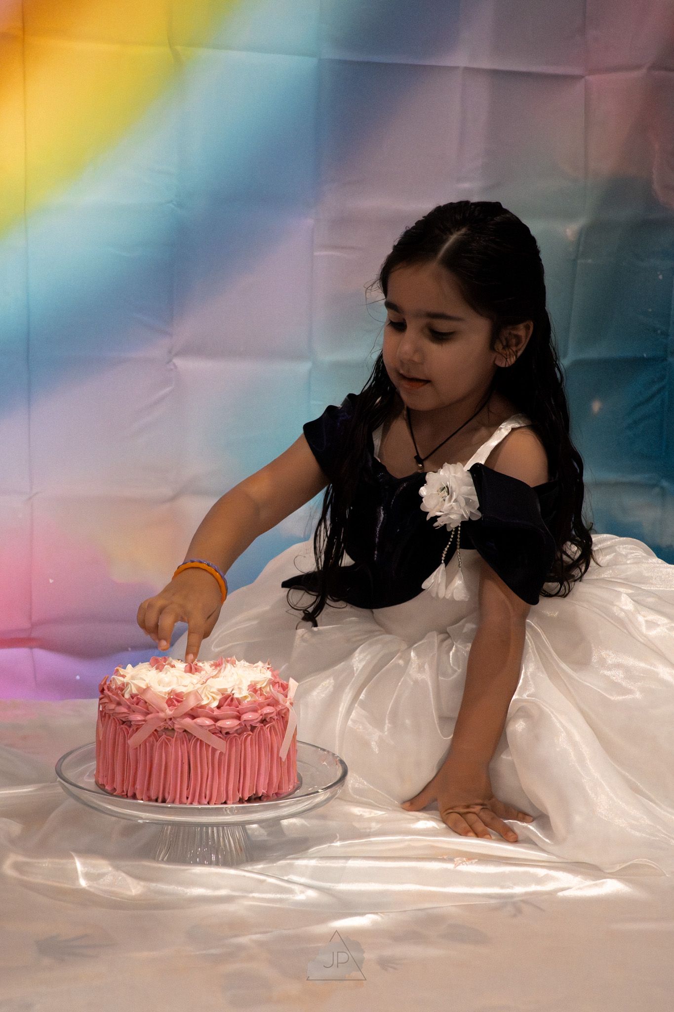 A young girl in a white dress reaches for a pink birthday cake against a colorful rainbow backdrop.