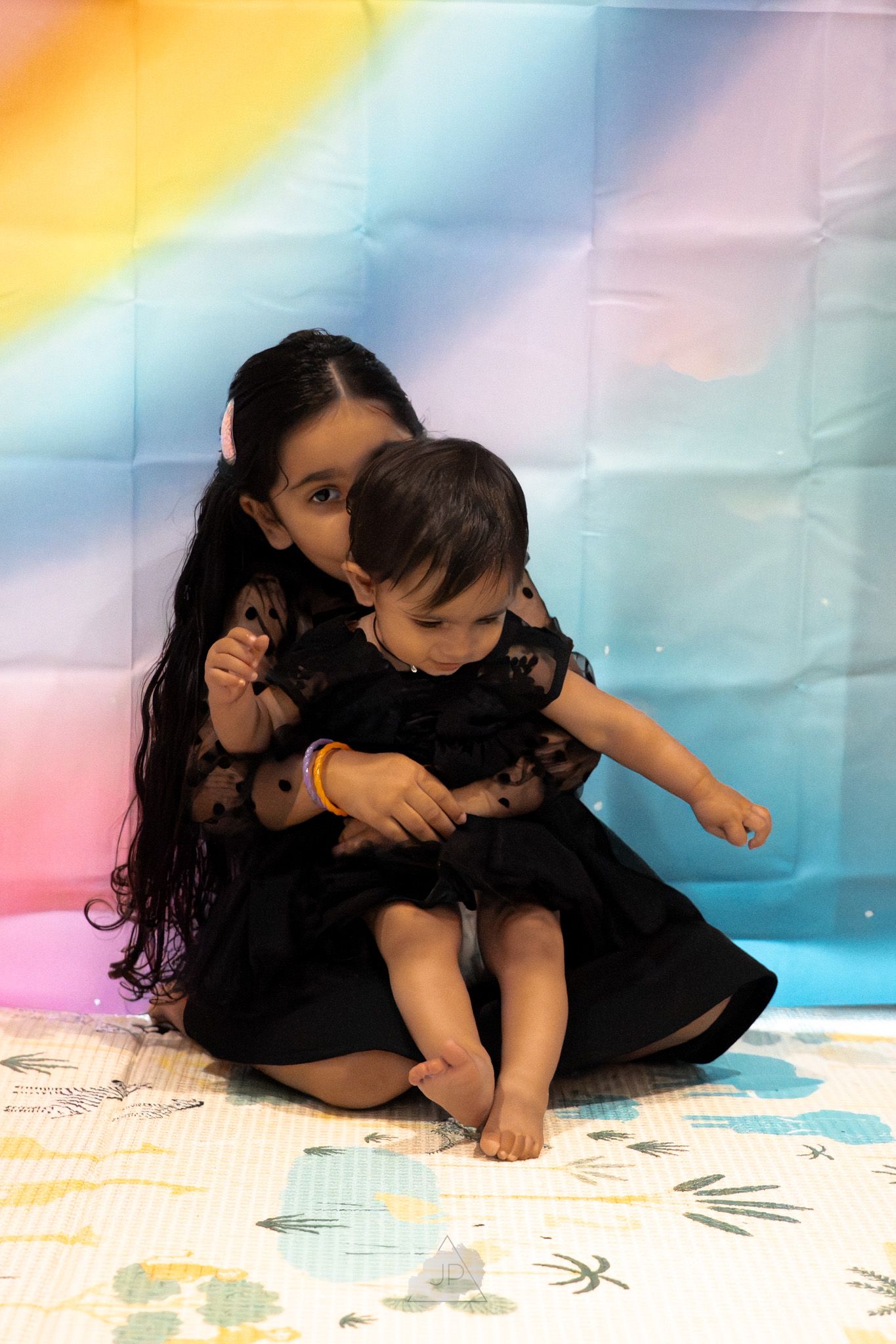 Two children in black outfits play together on a patterned floor mat with colorful lighting in the background.